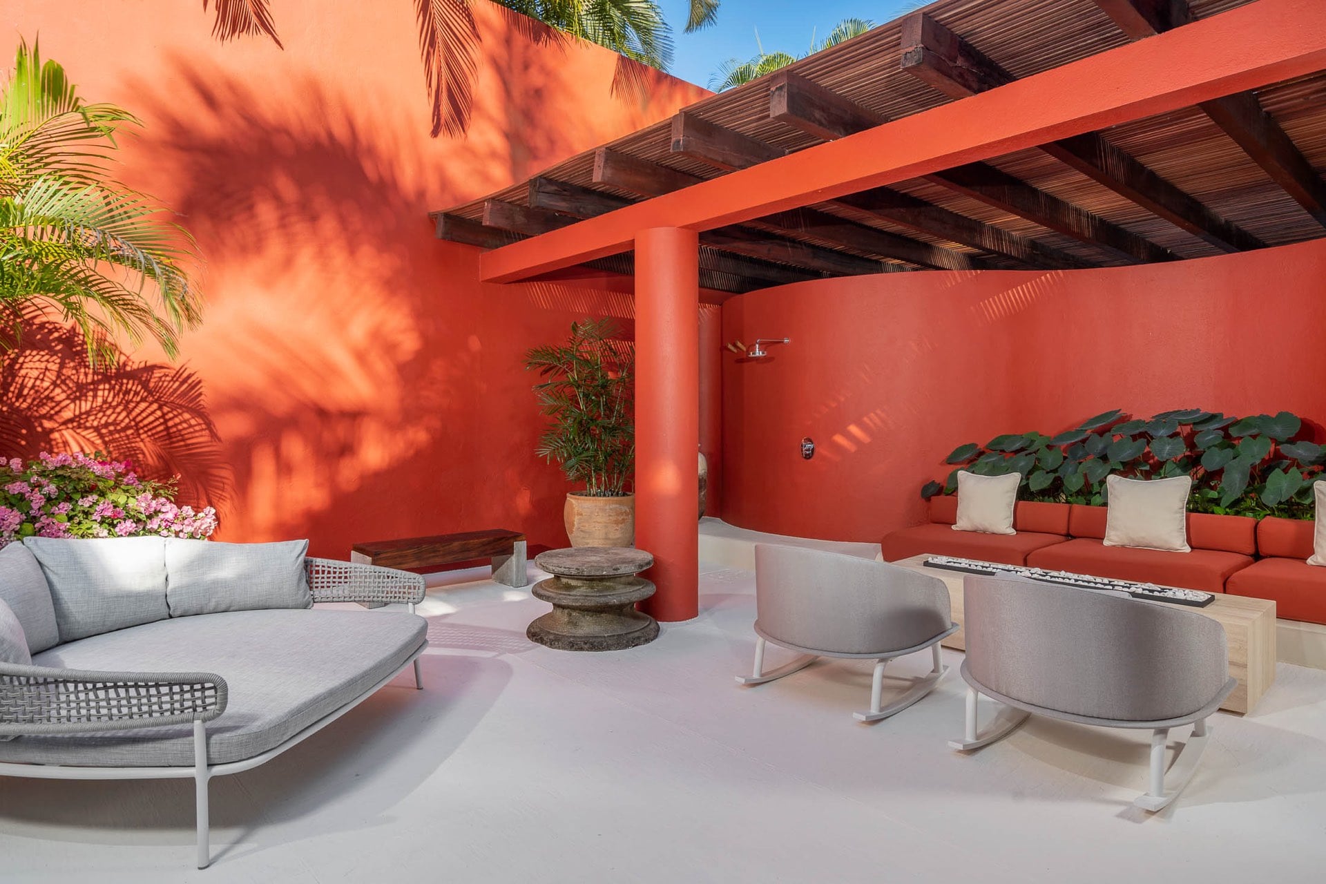 Covered outdoor terrace with coral-red stucco walls, slatted wood pergola, woven daybed, rocking lounge chairs, terracotta built-in sofa, elephant-ear planters, and outdoor shower, Villa Pacífica, Ranchos Estates, Punta Mita.