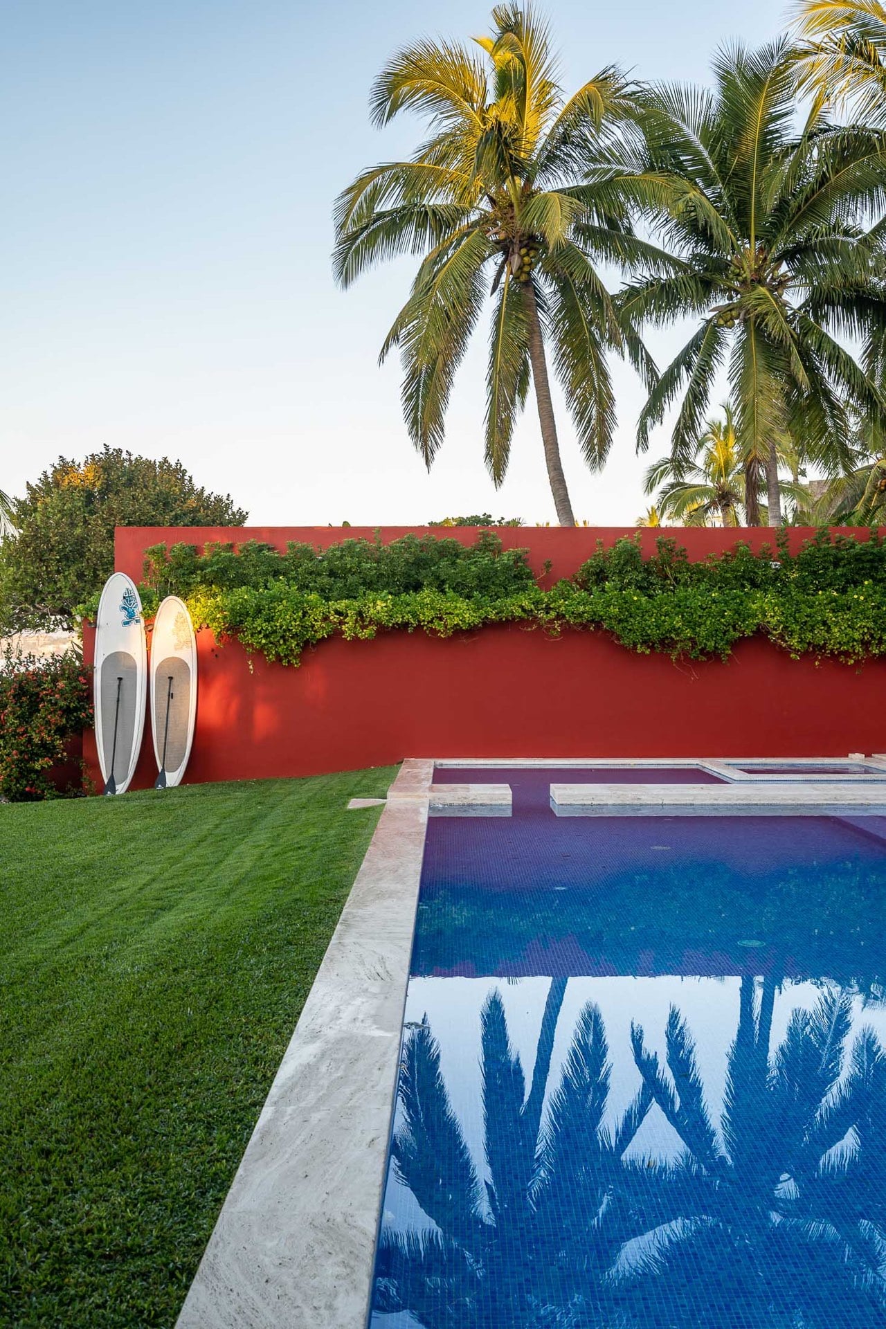 Pool edge at dusk with palm reflections on blue mosaic surface, green lawn, two standup paddleboards against coral-red garden wall, coconut palms overhead, Villa Pacífica, Ranchos Estates, Punta Mita.