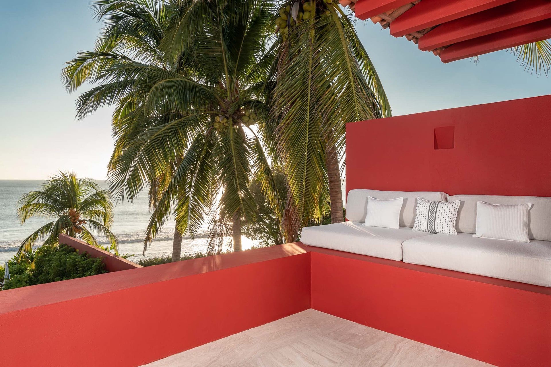 Upper terrace with white built-in sofa set against coral-red parapet wall, coconut palms framing a direct Pacific Ocean and beach view at golden hour, Villa Pacífica, Ranchos Estates, Punta Mita.