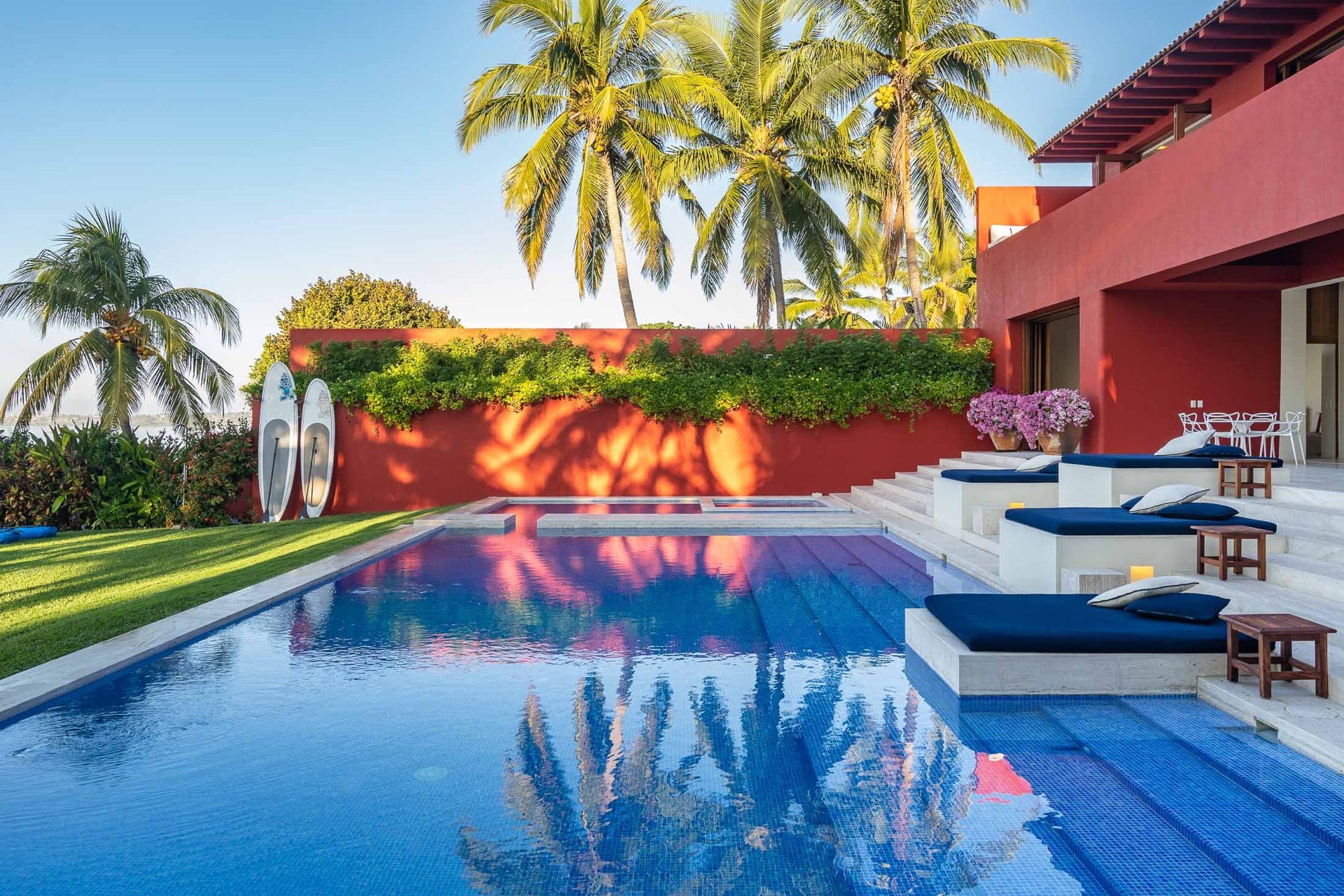 Wide view of rectangular pool reflecting coral-red villa facade and palms, navy chaise longues on stepped terrace, two paddleboards at garden wall, ocean at left horizon, Villa Pacífica, Ranchos Estates, Punta Mita.