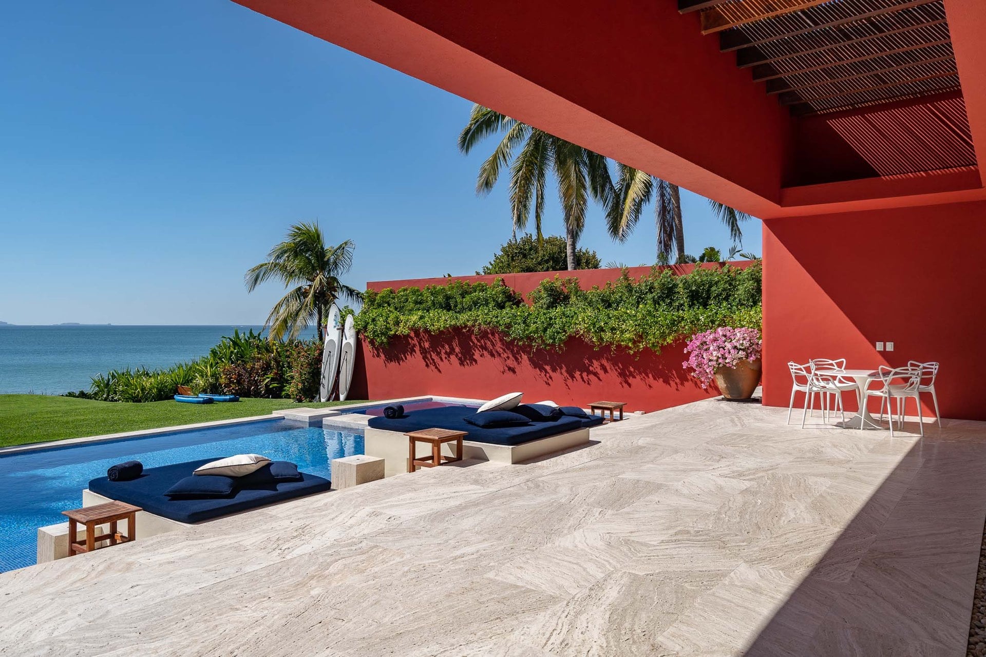 Travertine pool deck viewed from under pergola shade, navy sun loungers beside pool, coral-red walls, paddleboards at garden wall, Pacific horizon visible, white Masters chairs at outdoor dining table, Villa Pacífica, Ranchos Estates, Punta Mita.
