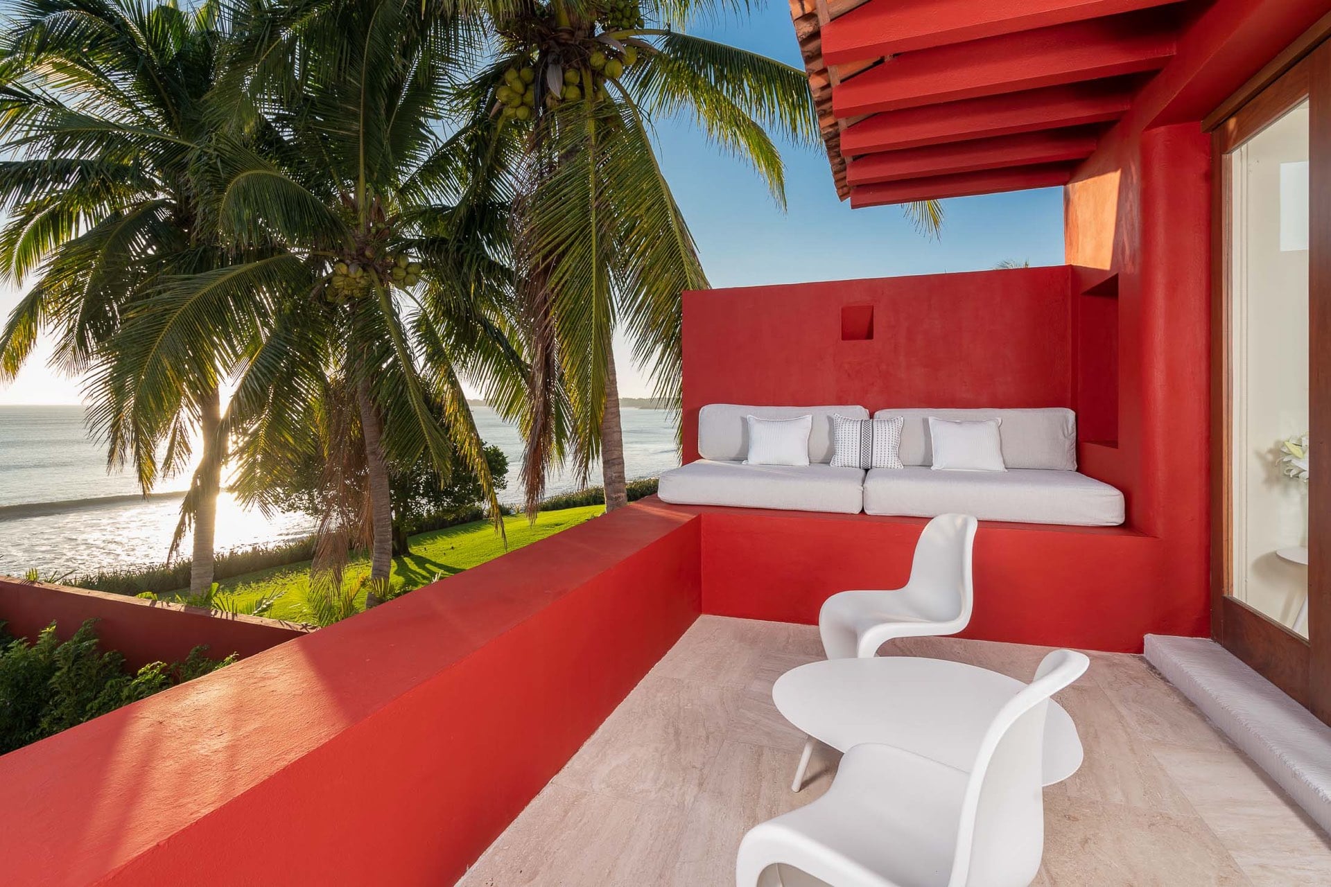 Private upper terrace with white built-in daybed and two Panton chairs, coral-red parapet walls, coconut palms and Pacific beach at sunset in direct sightline, Villa Pacífica, Ranchos Estates, Punta Mita.