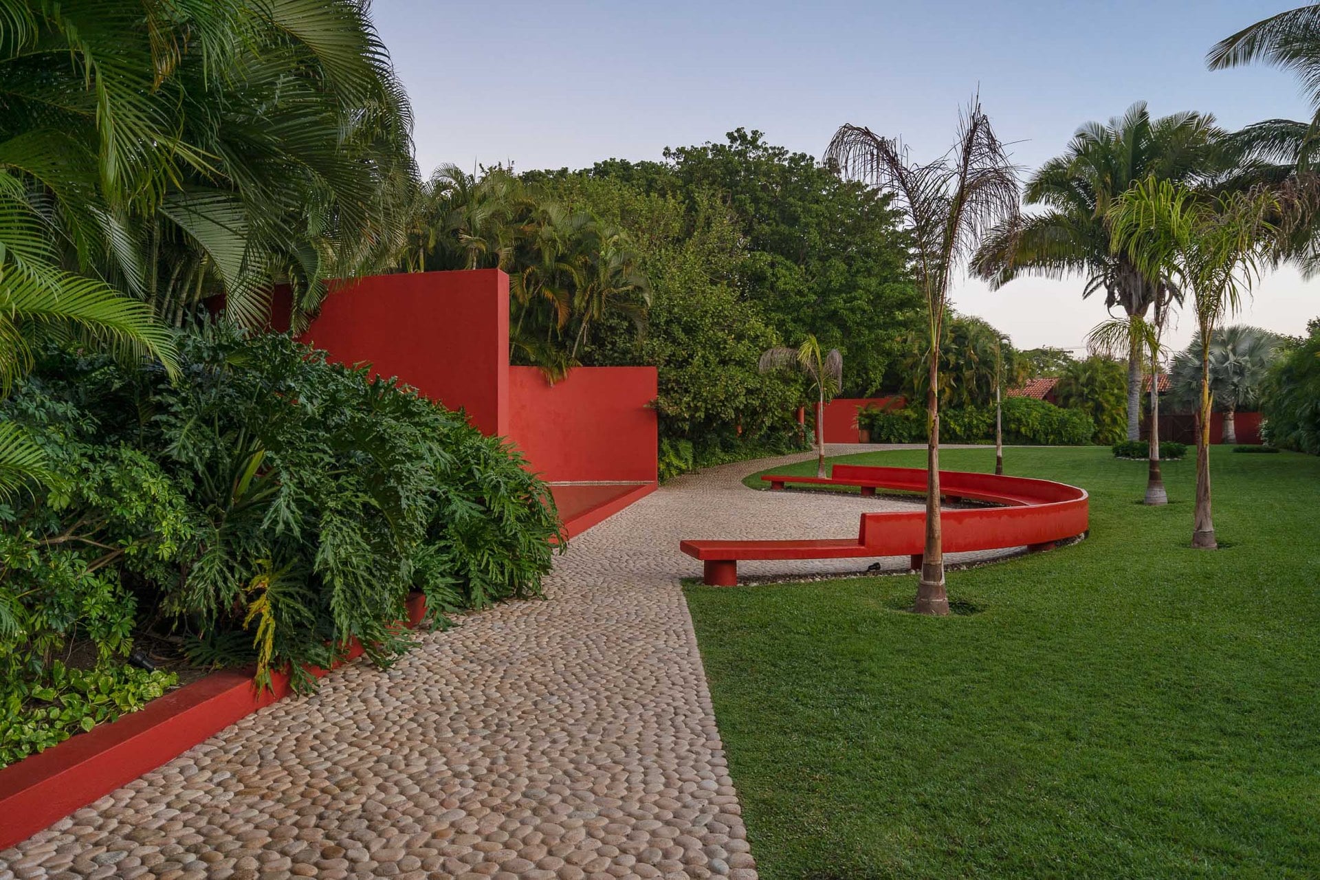 Cobblestone driveway winding through manicured lawn with coral-red curved garden walls and low benches, royal palms and dense tropical plantings at dusk, Villa Pacífica, Ranchos Estates, Punta Mita.