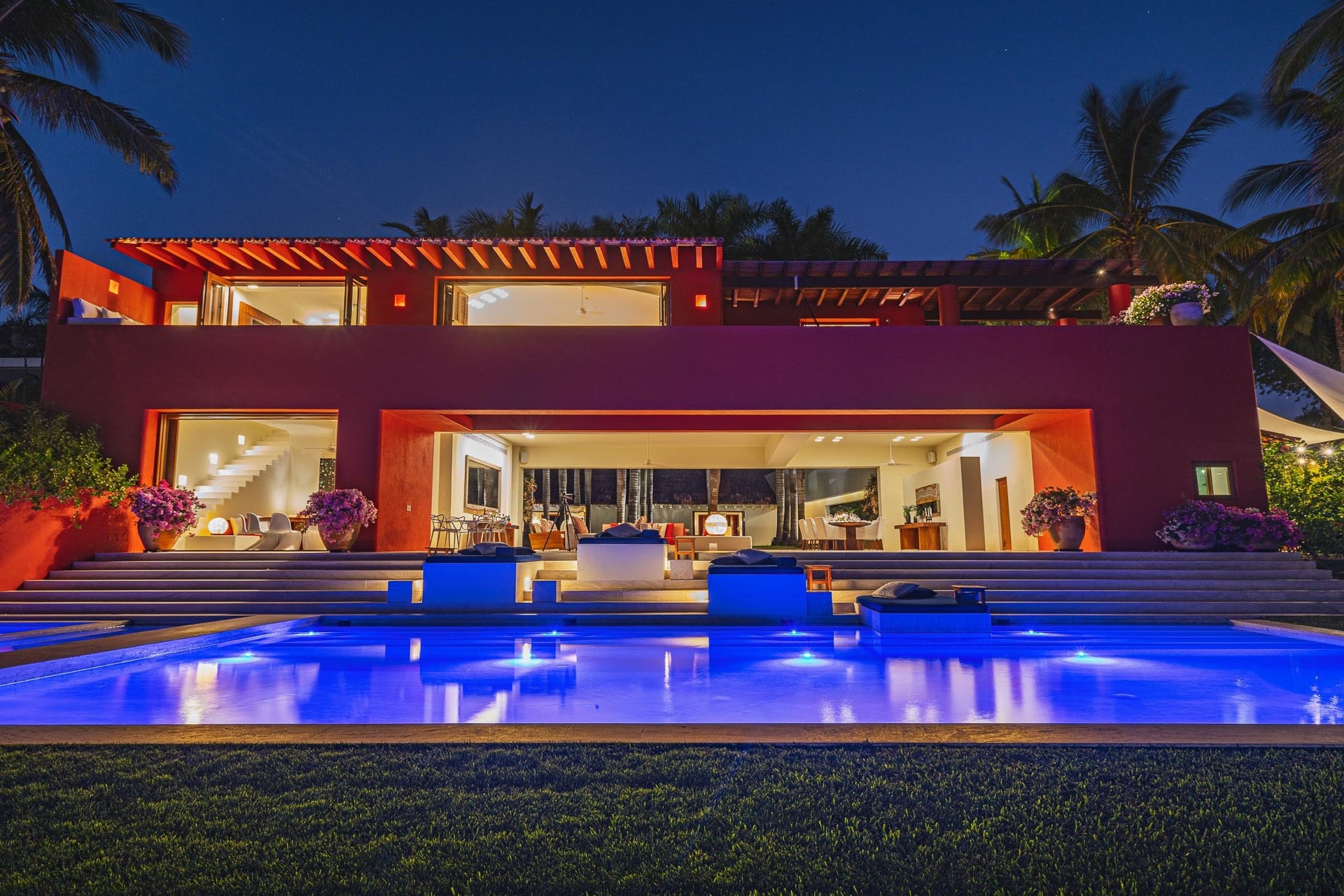 Villa exterior at night with pool lit electric blue in foreground, open ground-floor living pavilion glowing warm amber, upper terrace with red pergola, bougainvillea urns flanking the entry steps, Villa Pacífica, Ranchos Estates, Punta Mita.