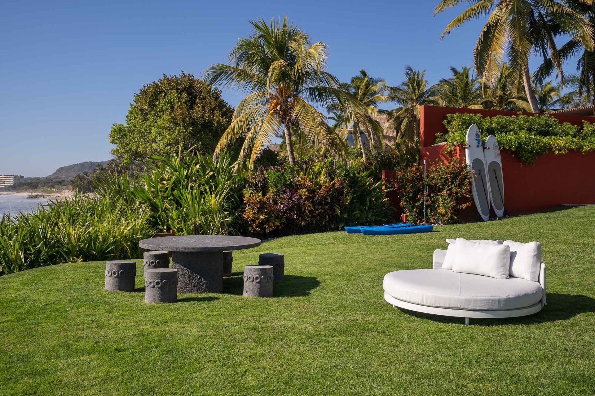 Beachfront lawn with white round daybed and volcanic-stone game table with four carved stools, paddleboards and a kayak at garden wall, Punta Mita bay visible at left, Villa Pacífica, Ranchos Estates, Punta Mita.