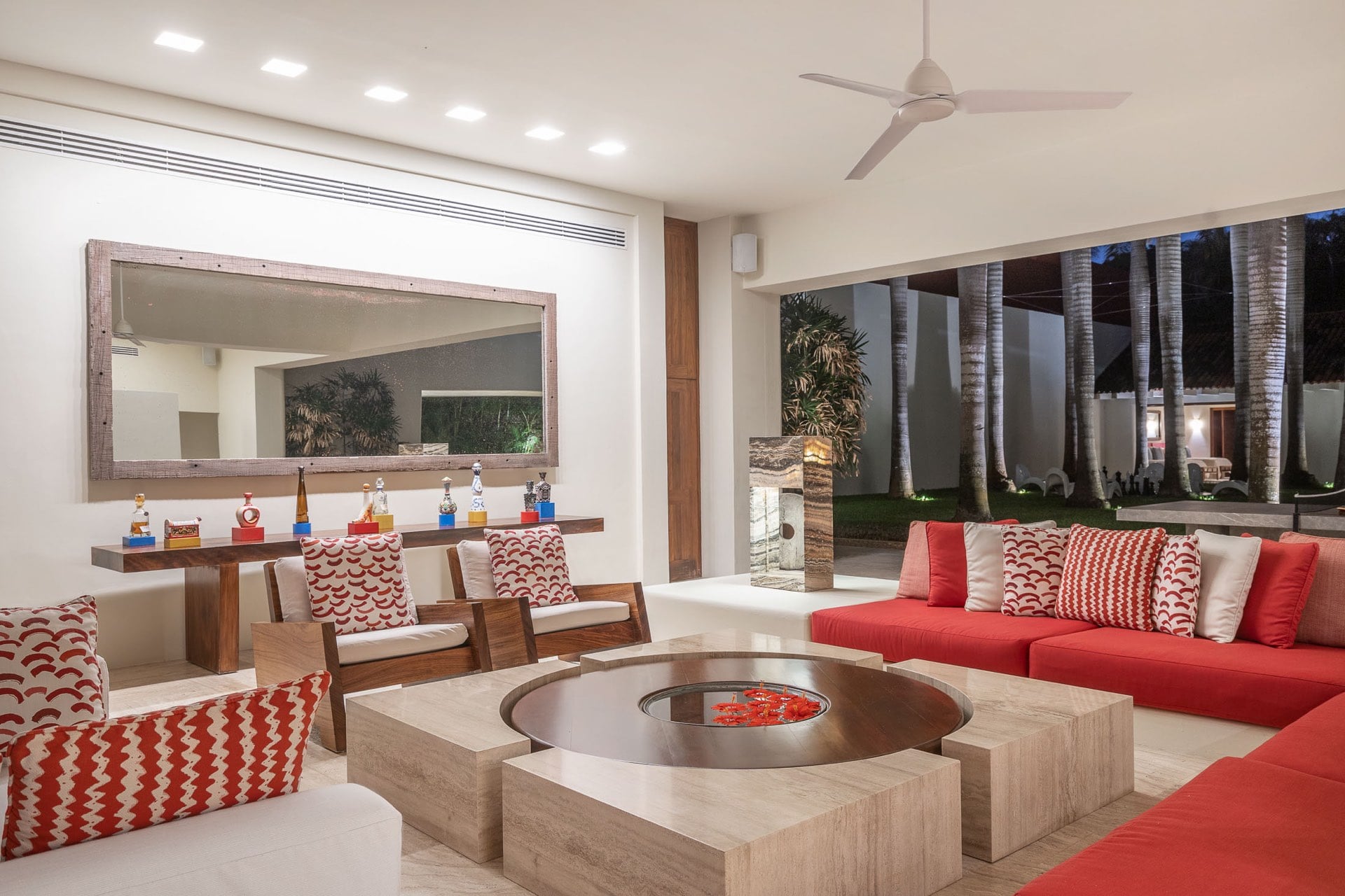 Open-air living pavilion at night with red sectional sofas, graphic-print cushions, round travertine fire-pit coffee table, walnut armchairs, and a tequila bottle display on a floating shelf below a large mirror, Villa Pacífica, Ranchos Estates, Punta Mita.