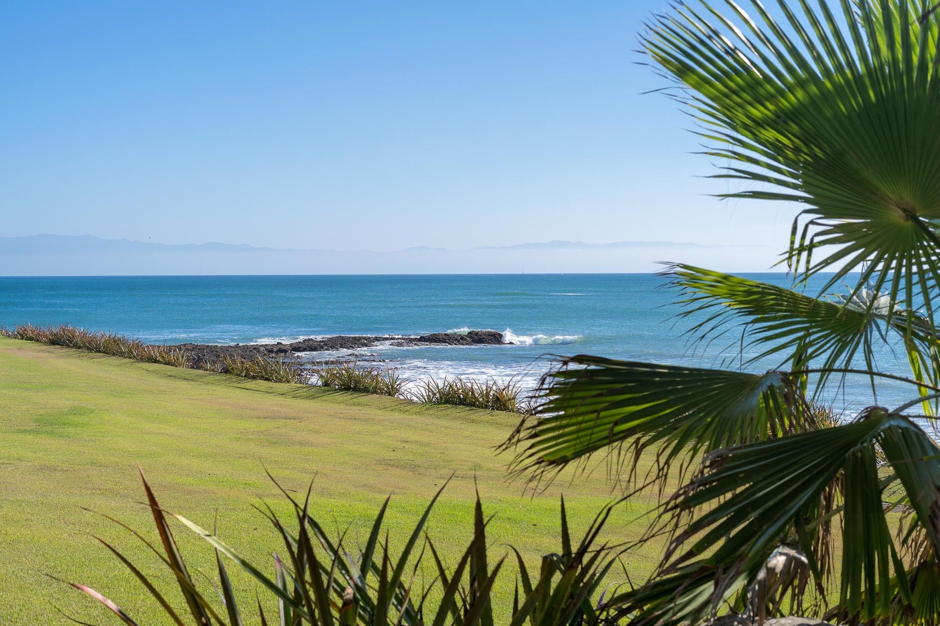 Banderas Bay seen through a fringe of palm fronds above a close-cropped coastal lawn, Sierra Madre silhouette on the horizon, Villa Pacífica, Ranchos Estates, Punta Mita.