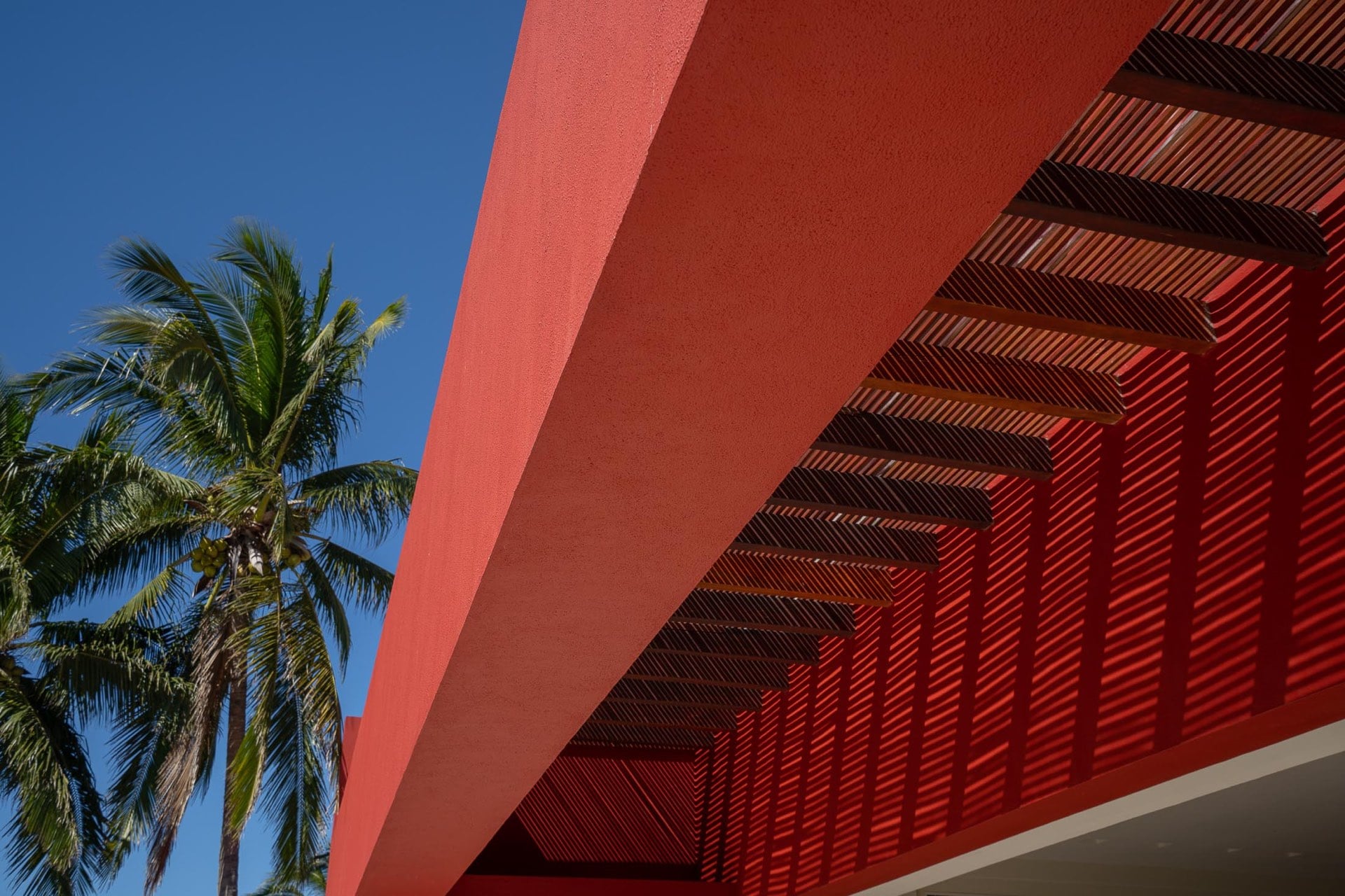 Bold red stucco eave with a slatted pergola detail photographed from below, a coconut palm rising against clear blue sky, Villa Pacífica, Ranchos Estates, Punta Mita.