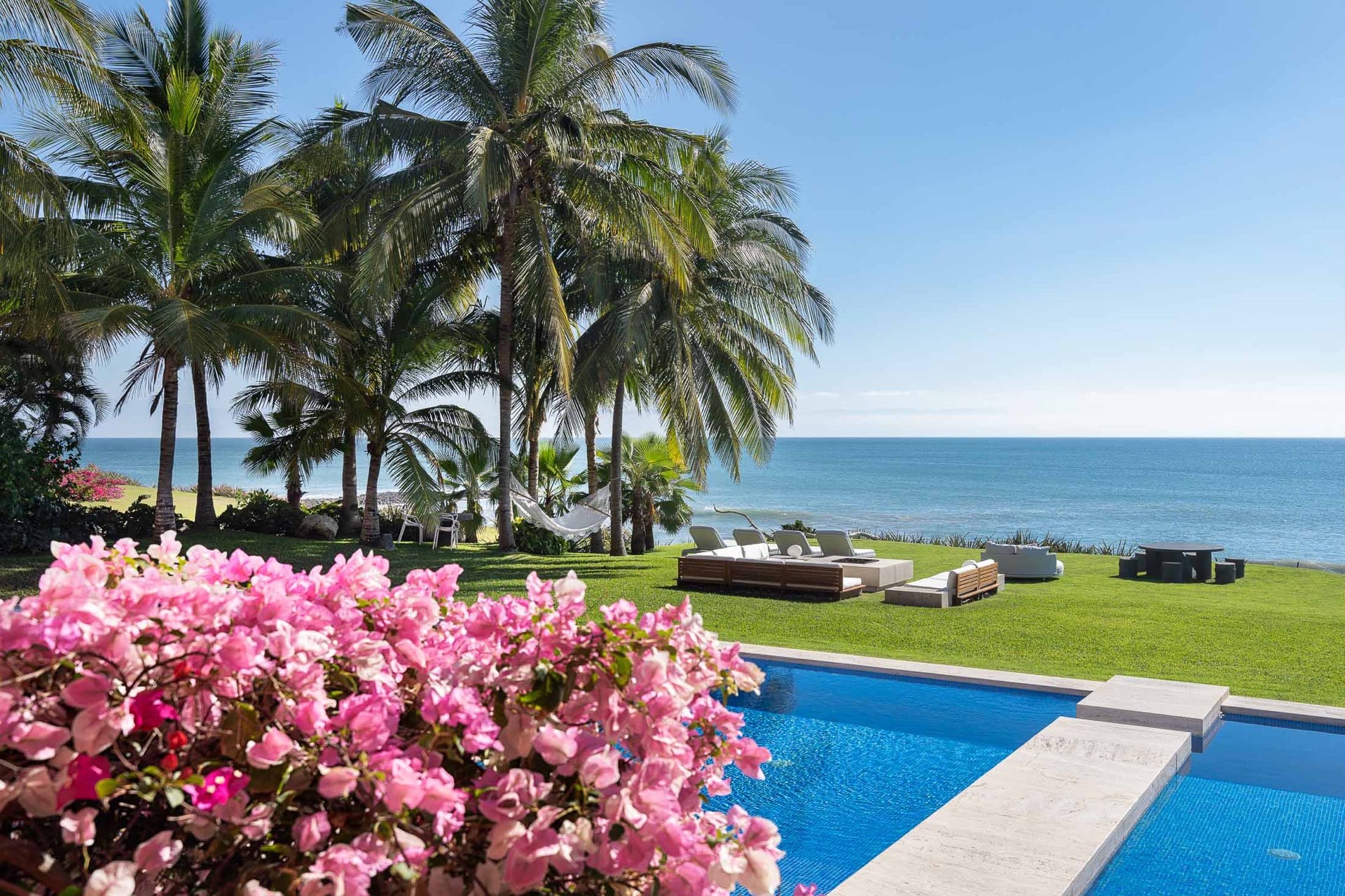 Pool edge bordered by pink bougainvillea in bloom, sun loungers on the beachfront lawn, coconut palms, and Banderas Bay beyond, Villa Pacífica, Ranchos Estates, Punta Mita.
