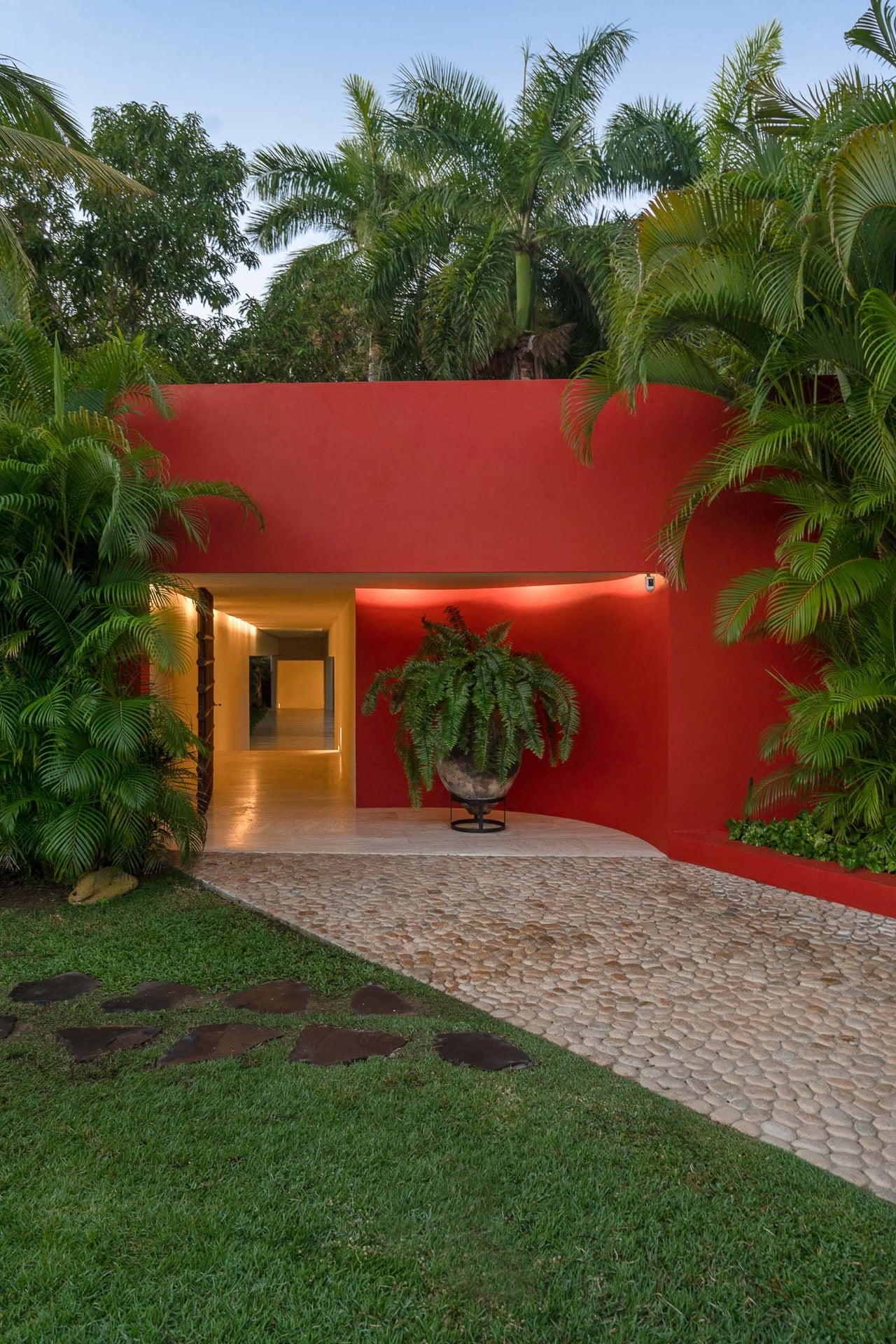 Villa entrance portal in red stucco with a lit passageway, stone cobble forecourt, large potted fern, and tropical palms overhead, Villa Pacífica, Ranchos Estates, Punta Mita.