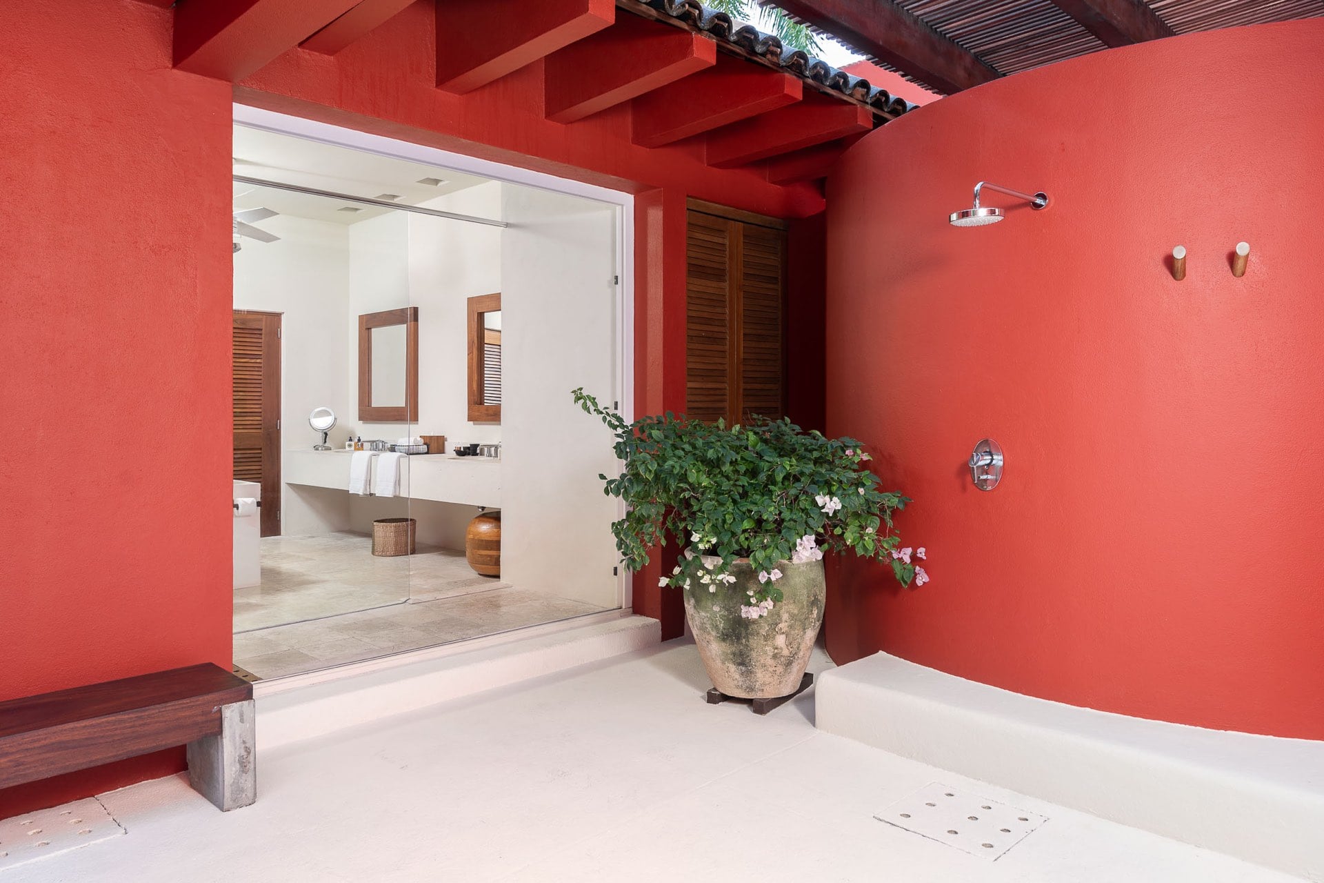 Open-air shower alcove on a red stucco courtyard wall with rain head and robe hooks, antique clay pot with flowering plant, glass door to the interior bath, Villa Pacífica, Ranchos Estates, Punta Mita.