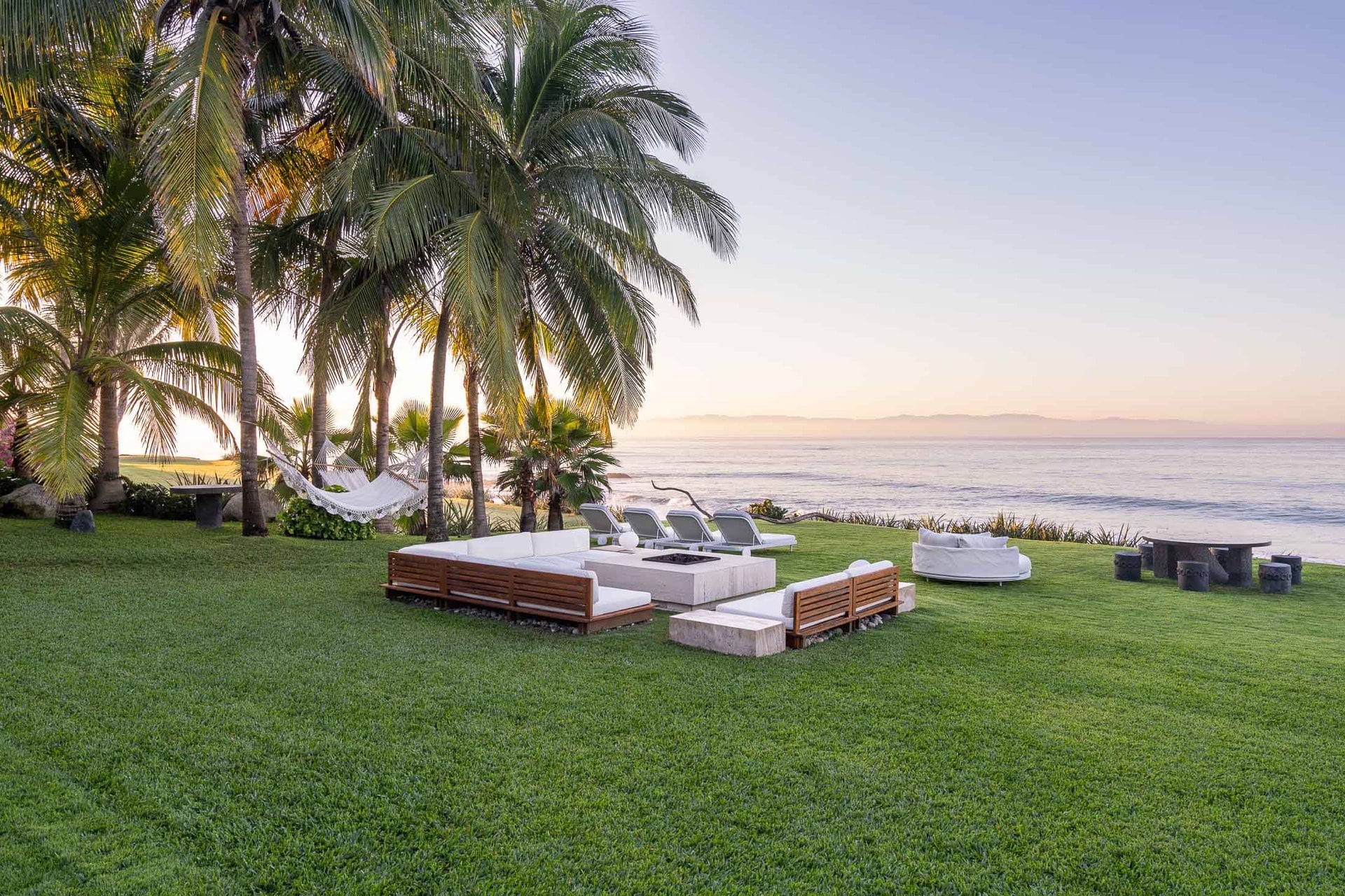 Sunken beachfront lounge with a fire pit and white cushioned seating on the lawn at dusk, hammocks between coconut palms, Banderas Bay glowing at the horizon, Villa Pacífica, Ranchos Estates, Punta Mita.
