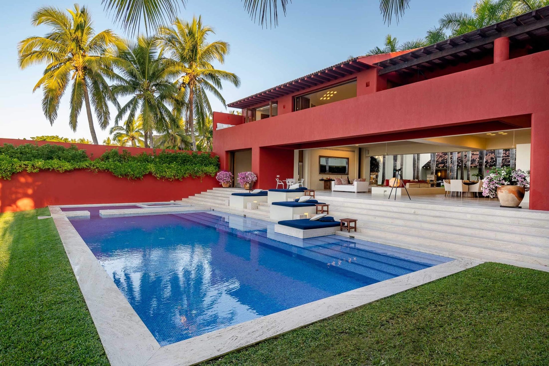 Rectangular pool with in-water sun shelves and navy cushions at dusk, travertine steps leading to a lit living pavilion behind the red stucco facade, Villa Pacífica, Ranchos Estates, Punta Mita.