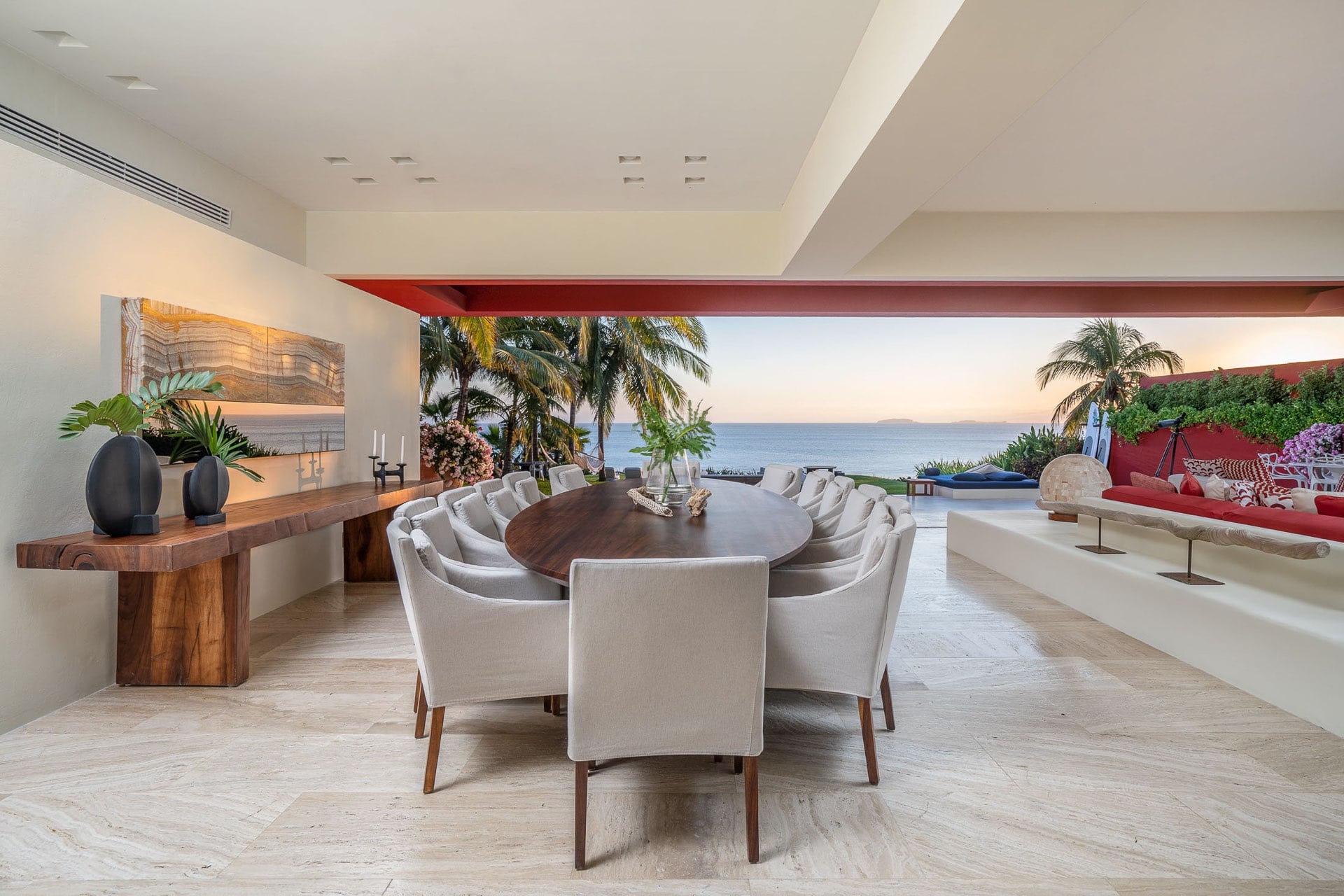 Open-air dining pavilion with an oval walnut table set for ten, a live-edge console with ceramic vessels, backlit onyx wall panel, and Banderas Bay at sunset framed by the open facade, Villa Pacífica, Ranchos Estates, Punta Mita.