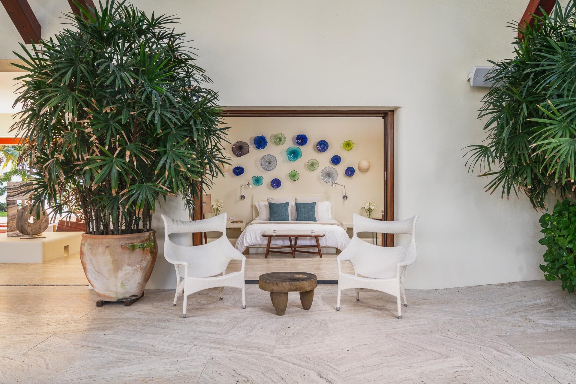 Two white bucket chairs and a carved wood stool in a sitting alcove framing a bedroom with Murano glass disc wall installation and white bedding, tall indoor palms flanking the niche, Villa Pacífica, Ranchos Estates, Punta Mita.