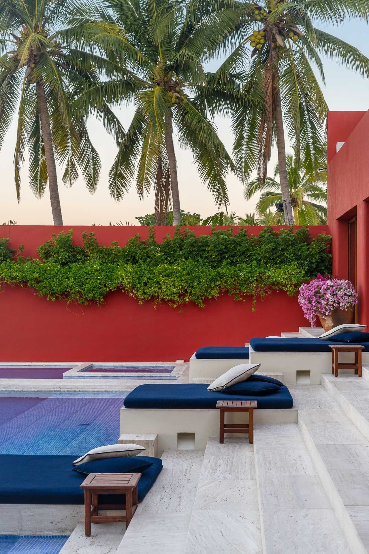 Navy sun loungers on travertine pool steps at dusk, creeping vine along the red perimeter wall, pink bougainvillea in a pot, coconut palms against a pale-blue sky, Villa Pacífica, Ranchos Estates, Punta Mita.