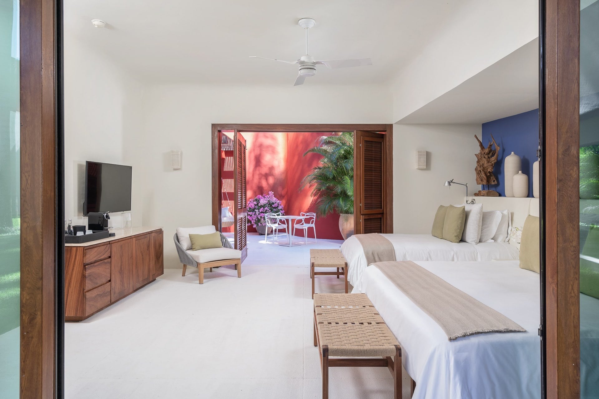 Guest room with two queen beds, walnut credenza with television, woven bench stools at the foot of each bed, blue accent wall, doors open to a private courtyard with bougainvillea, Villa Pacífica, Ranchos Estates, Punta Mita.