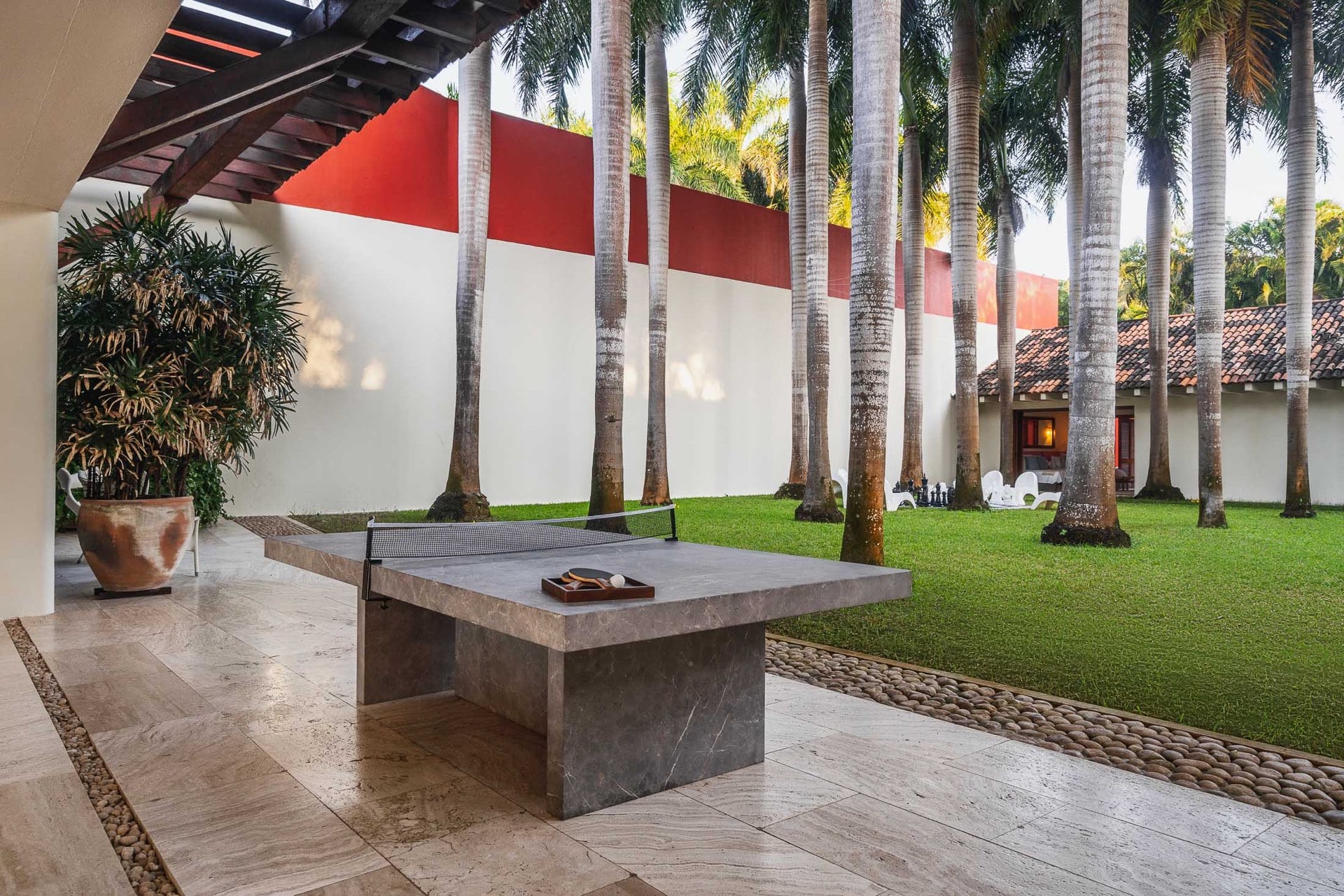 Slate ping-pong table on a covered travertine terrace, royal palms lining the manicured lawn, red-and-white compound wall behind, Villa Pacífica, Ranchos Estates, Punta Mita.