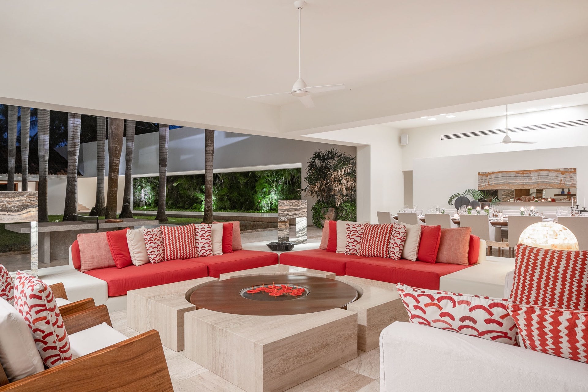 Main living pavilion at night showing a U-shaped red sectional with geometric patterned pillows, round walnut coffee table, and floodlit garden palms visible through the open facade, Villa Pacífica, Ranchos Estates, Punta Mita.