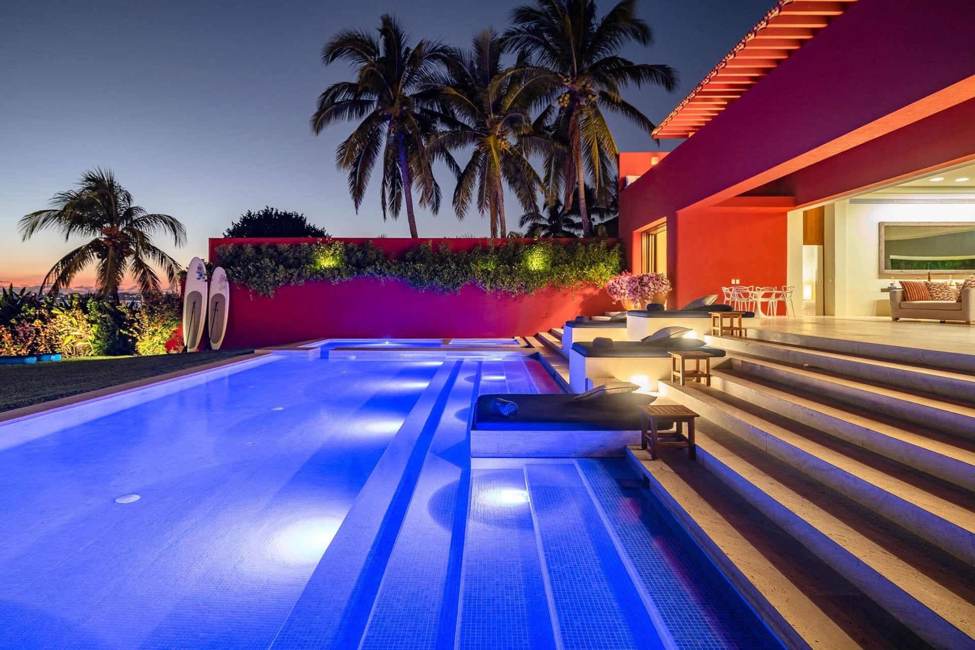 Illuminated pool at twilight with in-water sun shelves, travertine steps glowing amber, stand-up paddleboards propped against the red stucco wall, coconut palms silhouetted, Villa Pacífica, Ranchos Estates, Punta Mita.