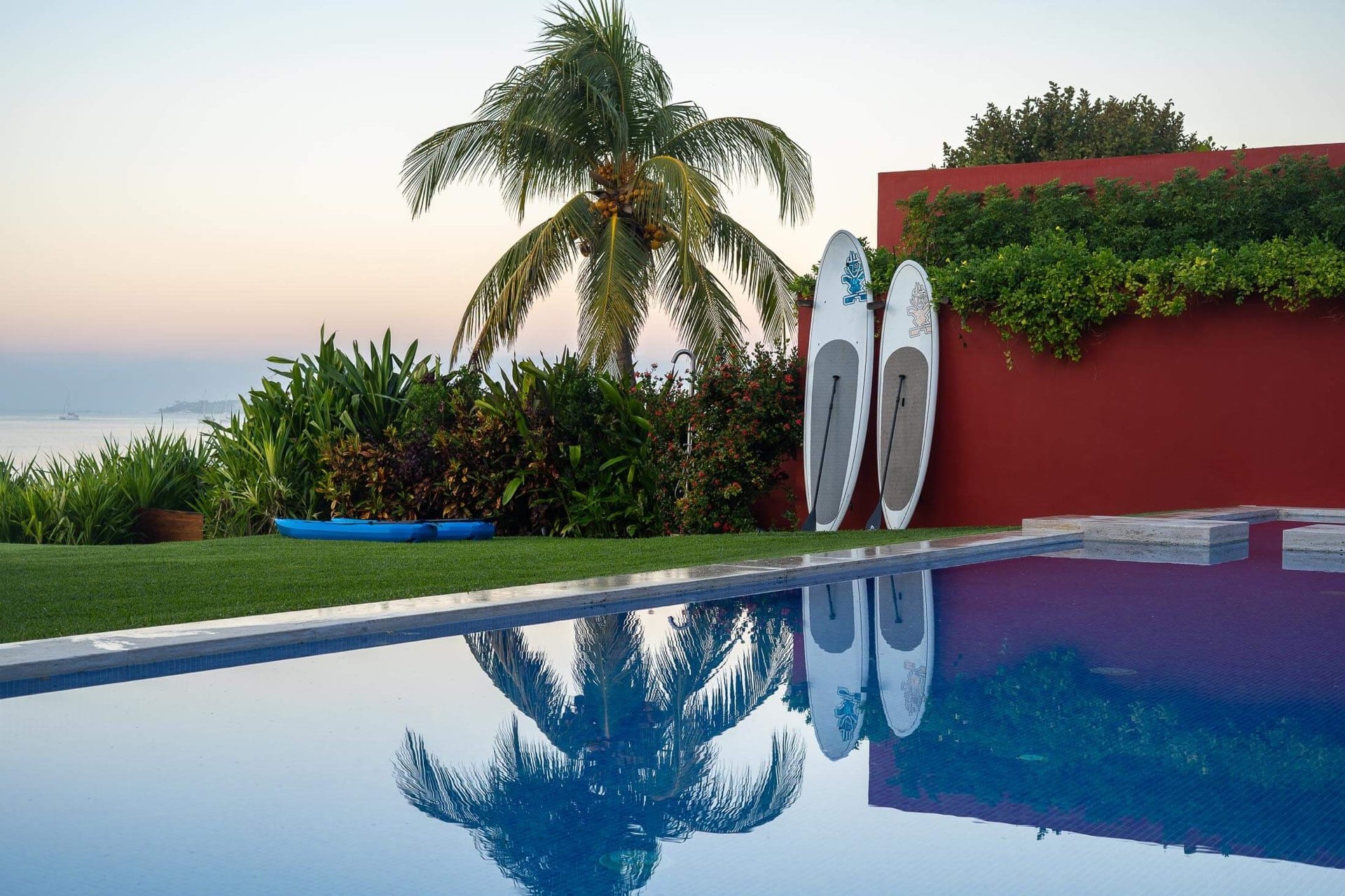 Still pool surface mirroring a coconut palm and two stand-up paddleboards leaned against the red perimeter wall, a kayak on the grass, the bay softly lit at dusk, Villa Pacífica, Ranchos Estates, Punta Mita.