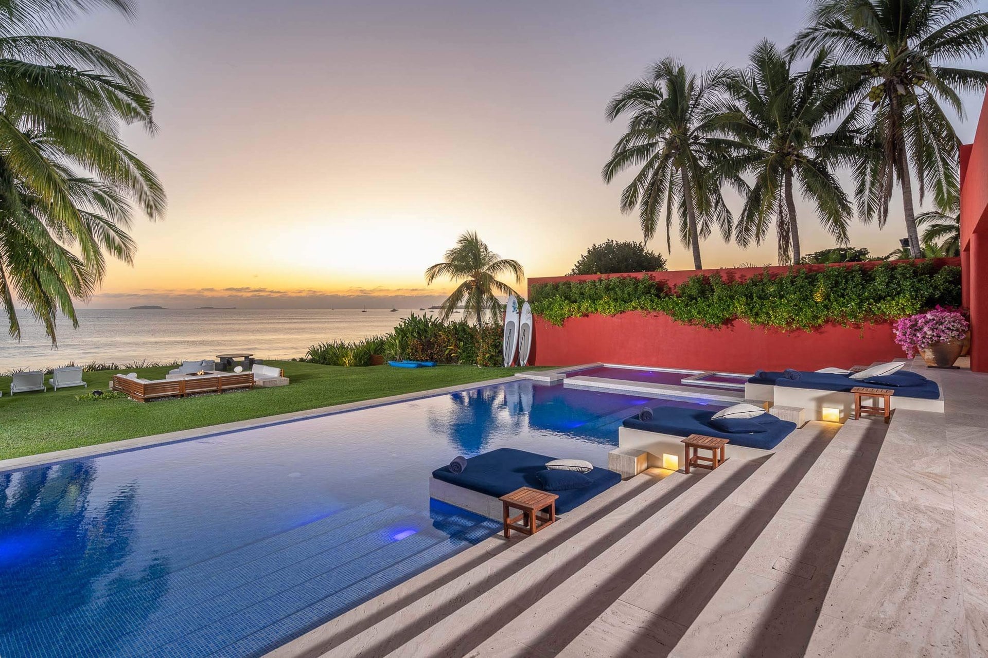 Wide pool panorama at sunset with in-water loungers and navy cushions on travertine coping, beachfront fire-pit seating and palms beyond, Banderas Bay horizon glowing orange, Villa Pacífica, Ranchos Estates, Punta Mita.