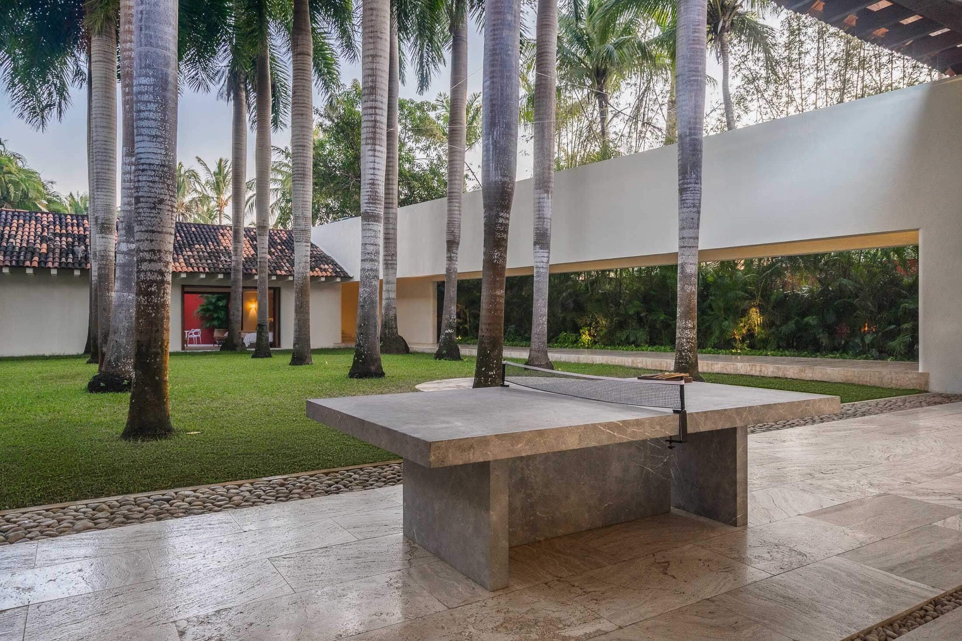Slate ping-pong table on a travertine court seen from a wide angle, royal palms bordering the lawn, white perimeter wall with a long horizontal window strip, guest casita visible at the far end, Villa Pacífica, Ranchos Estates, Punta Mita.