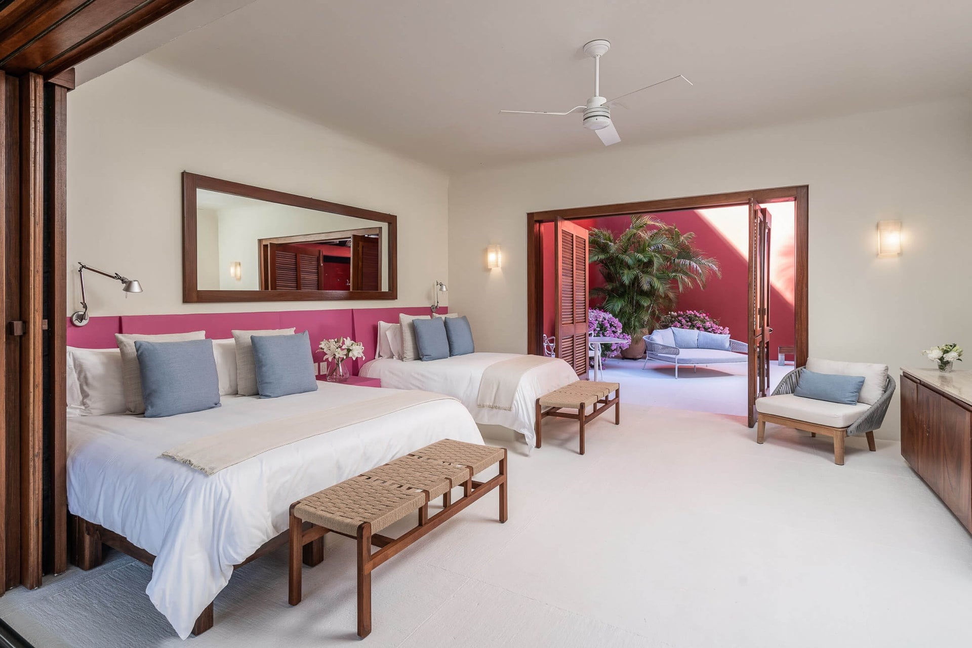 Guest room with two queen beds against a pink-painted headboard wall, wood-framed mirror above, rope-woven bench stools at the foot, folding doors open to a private red-walled courtyard with bougainvillea, Villa Pacífica, Ranchos Estates, Punta Mita.