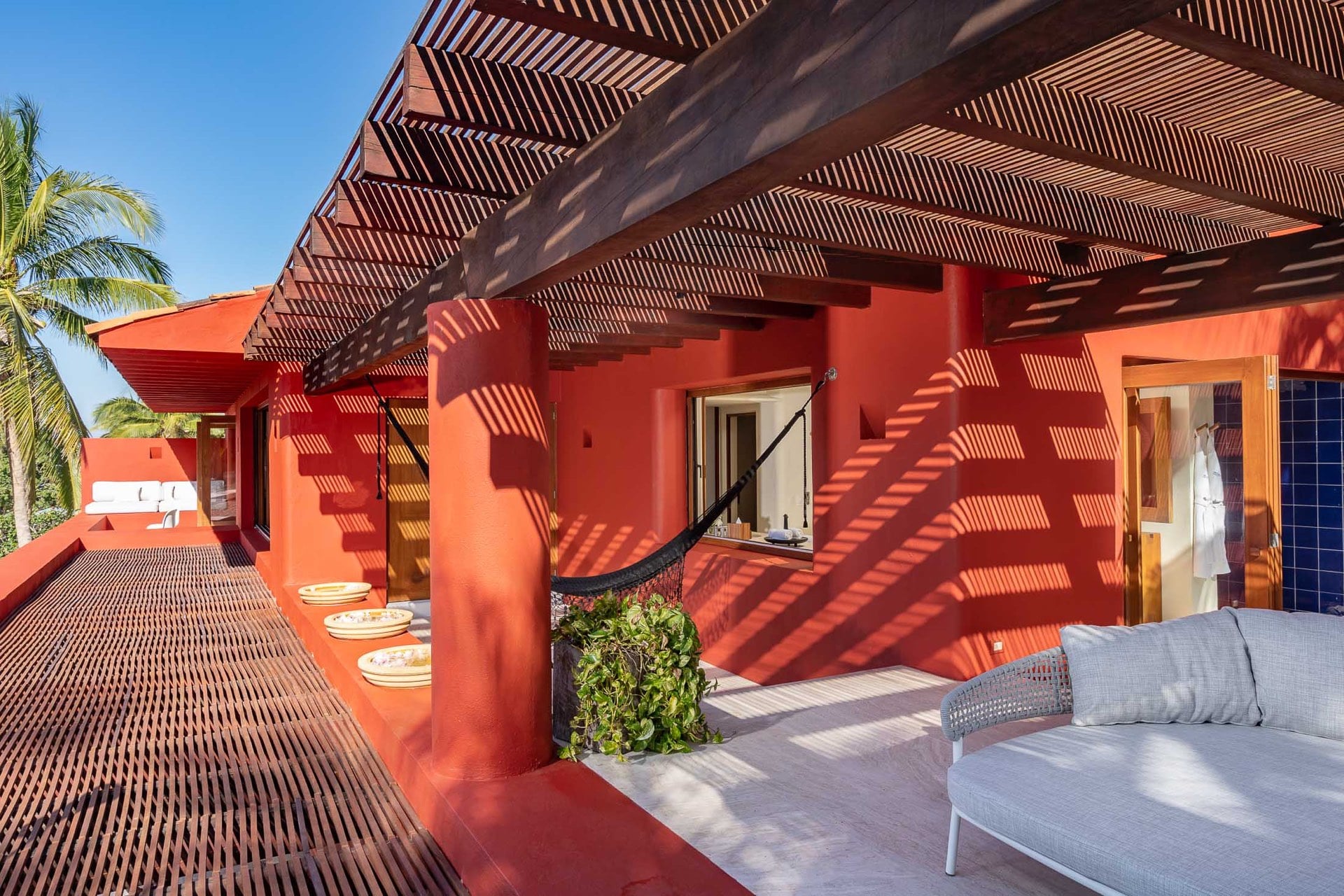 Upper rooftop terrace with a slatted pergola casting lattice shadows across red stucco walls, a hammock, an outdoor sofa, and a tiled sunning deck with clay platters, Villa Pacífica, Ranchos Estates, Punta Mita.