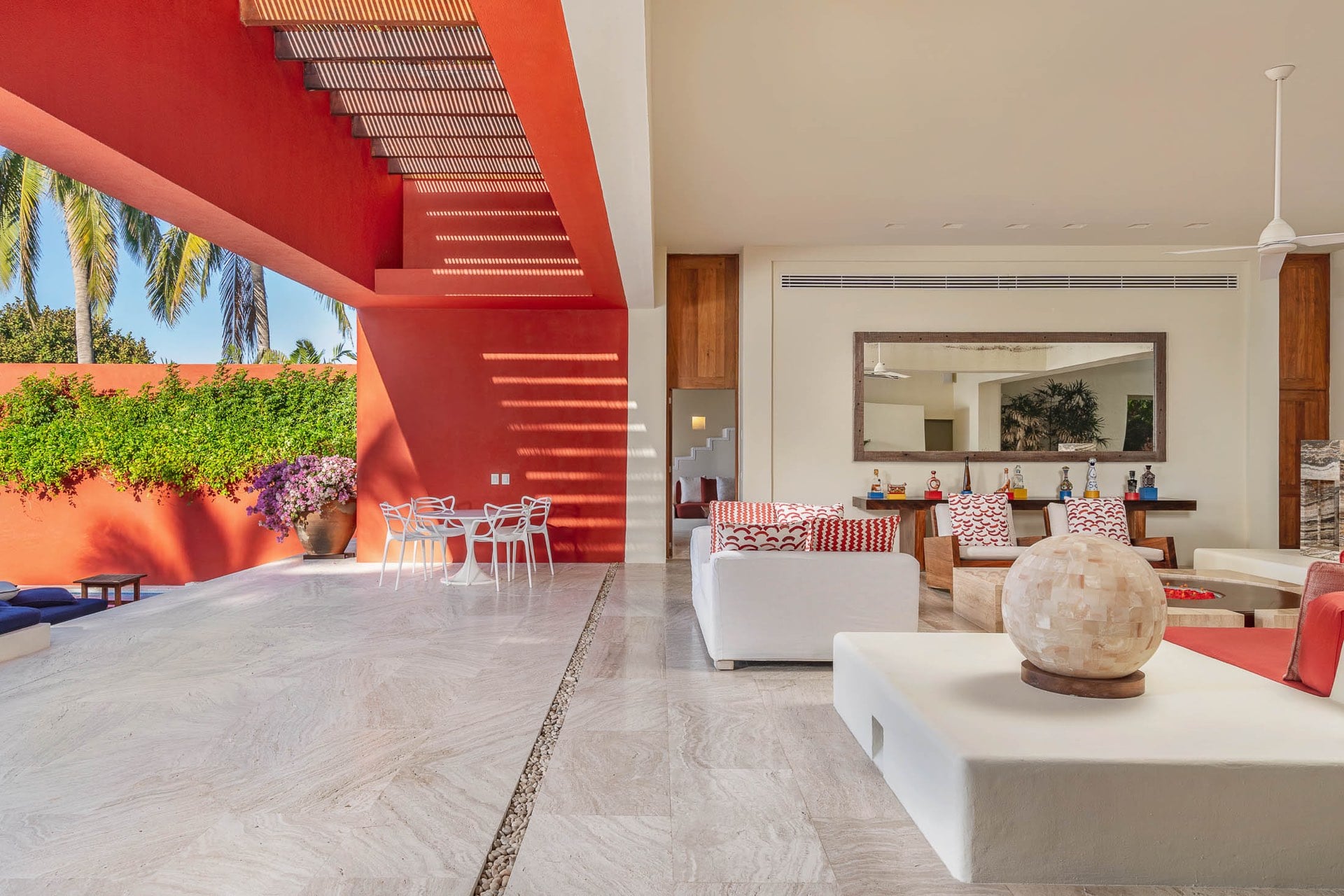 Travertine floor flowing from the interior living area with patterned red sofas and a folk-art bottle bar through a slatted red overhang to an outdoor bistro table in a bougainvillea courtyard, Villa Pacífica, Ranchos Estates, Punta Mita.