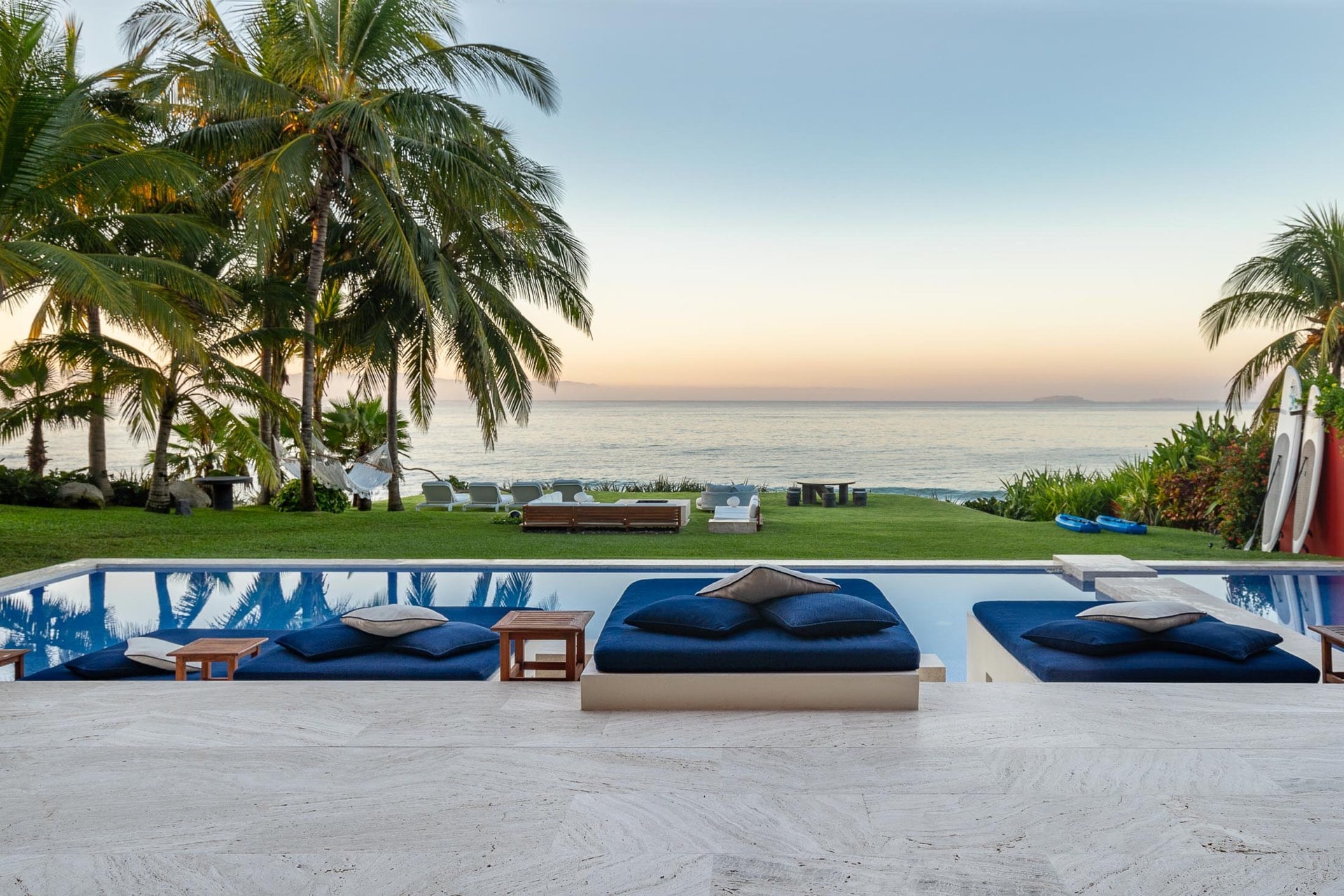Three navy in-water sun shelves on the pool deck at twilight, travertine coping in the foreground, beachfront lawn with hammocks and loungers beyond, Banderas Bay at a soft pastel dusk, Villa Pacífica, Ranchos Estates, Punta Mita.