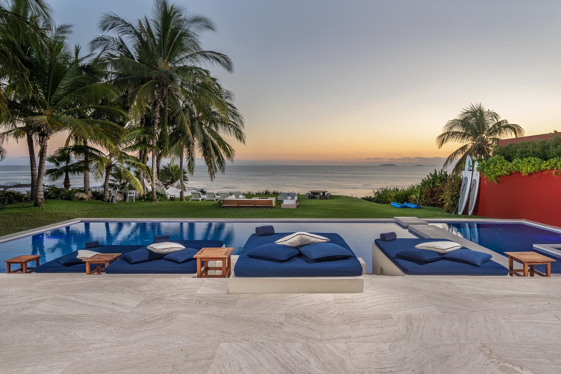 Four navy daybed loungers line the pool terrace at sunset, surfboards resting against the red perimeter wall while palm trees frame the Pacific beyond, Villa Pacífica, Ranchos Estates, Punta Mita.