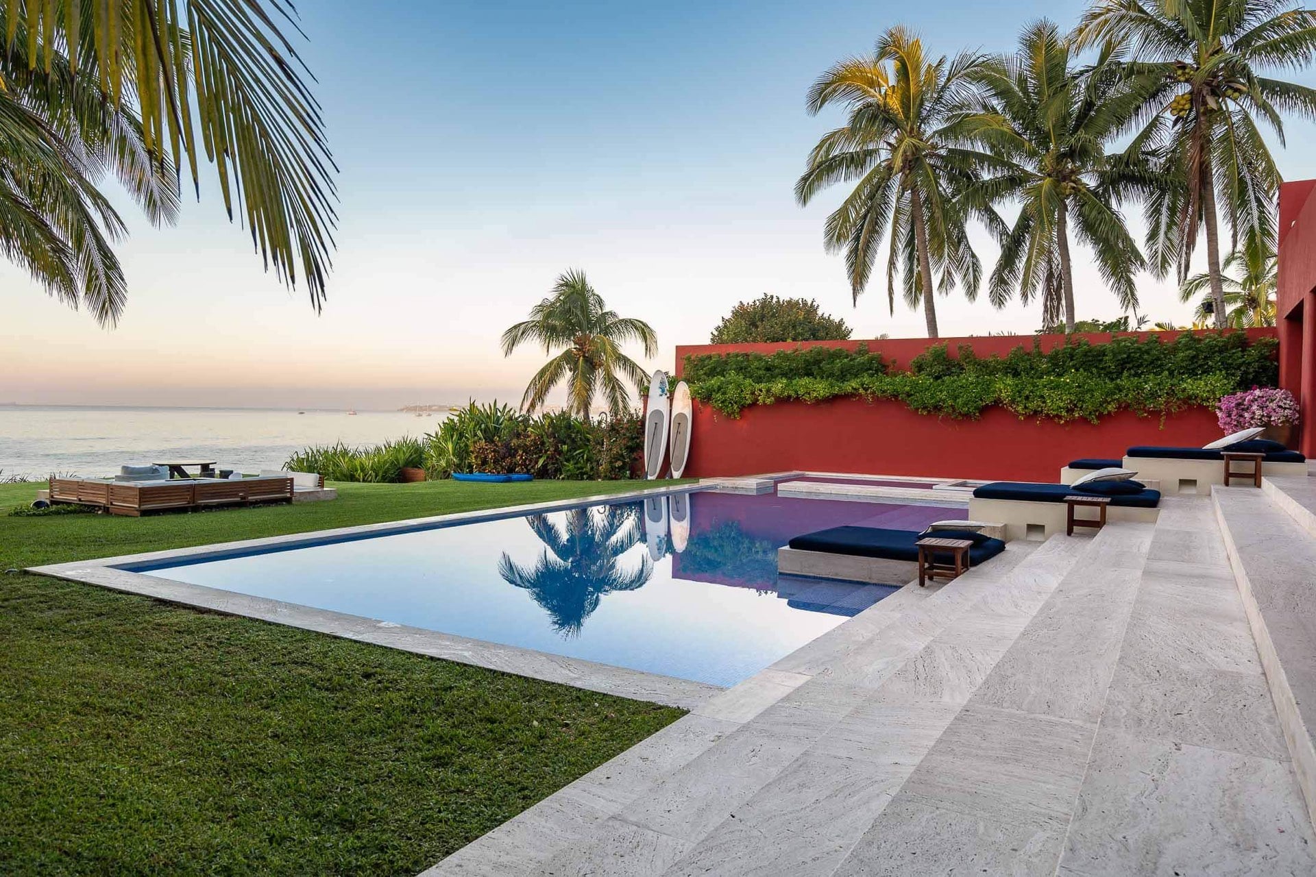 Dusk light catches the pool’s still surface as vines spill over the red garden wall and navy loungers sit ready at the far edge, Villa Pacífica, Ranchos Estates, Punta Mita.