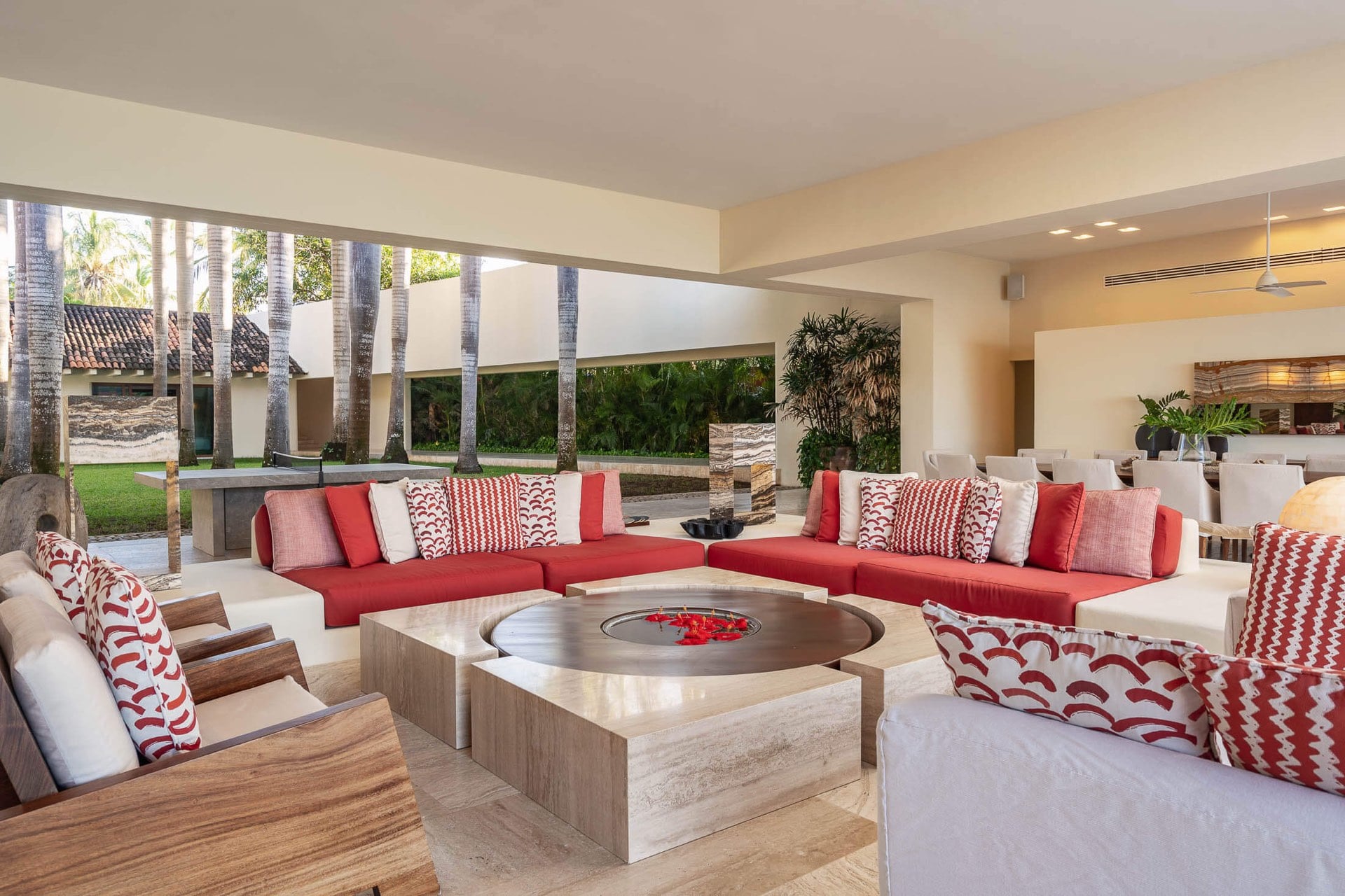 A large U-shaped red sectional wraps a travertine fire table in the open living pavilion, garden and pool visible through the frameless openings on three sides, Villa Pacífica, Ranchos Estates, Punta Mita.