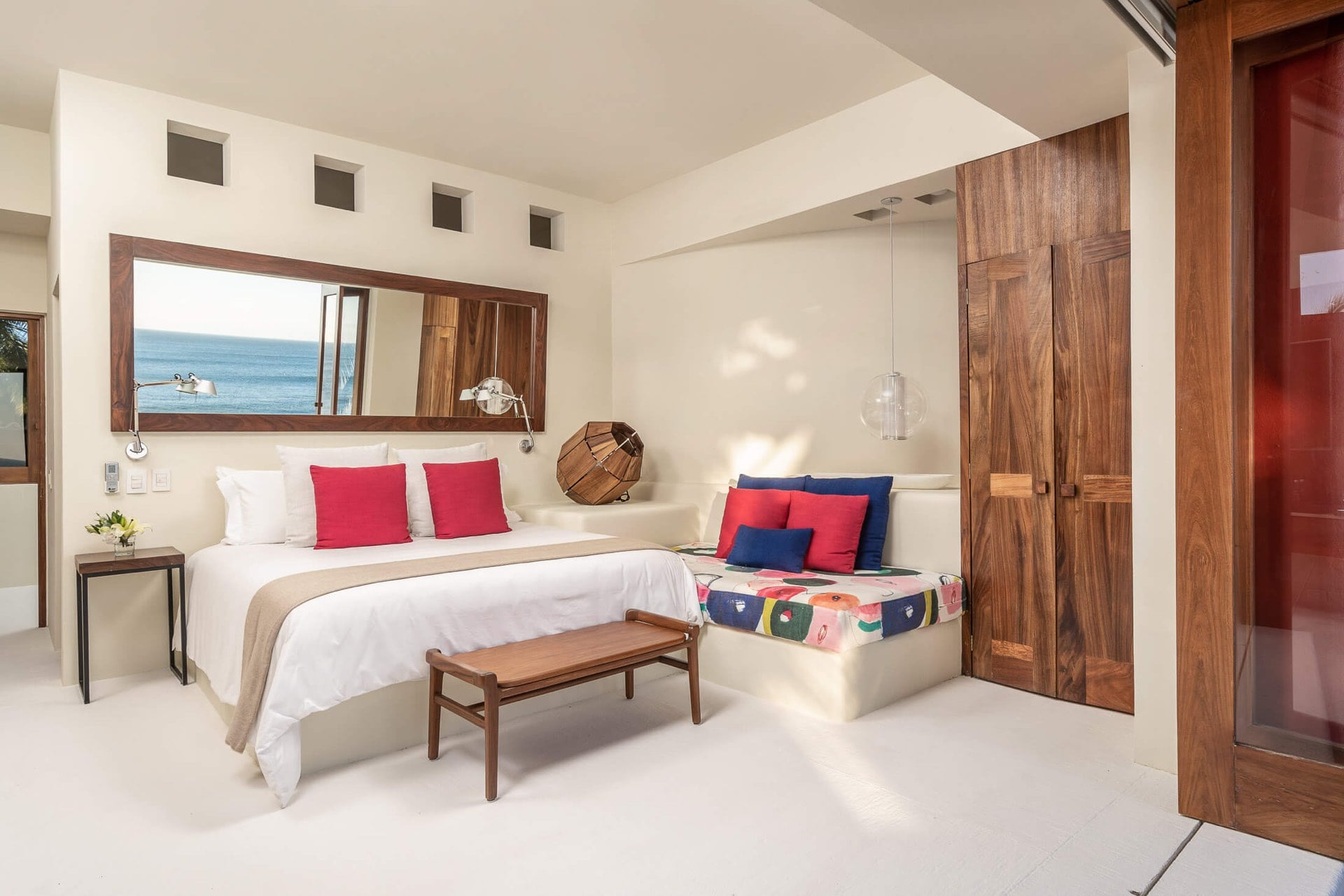 A king bed dressed in white faces a walnut-framed mirror that captures an ocean slice, while a reading nook with colorful cushions tucks beside the walnut entry door, Villa Pacífica, Ranchos Estates, Punta Mita.