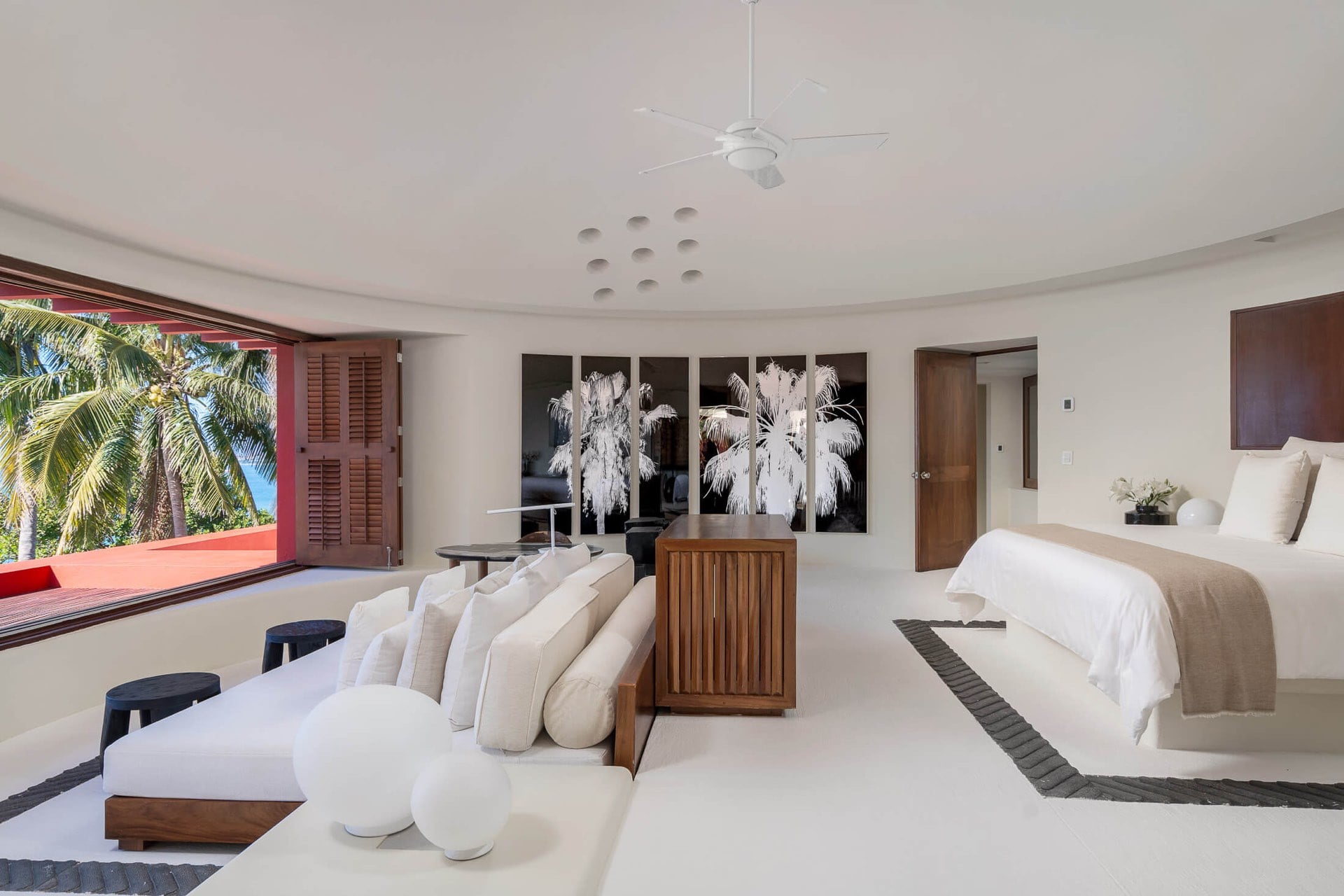 The primary suite’s curved ceiling spans a king bed in white and taupe, a walnut slatted chest, monochrome palm-print art panels, and shuttered doors left open to the palms outside, Villa Pacífica, Ranchos Estates, Punta Mita.