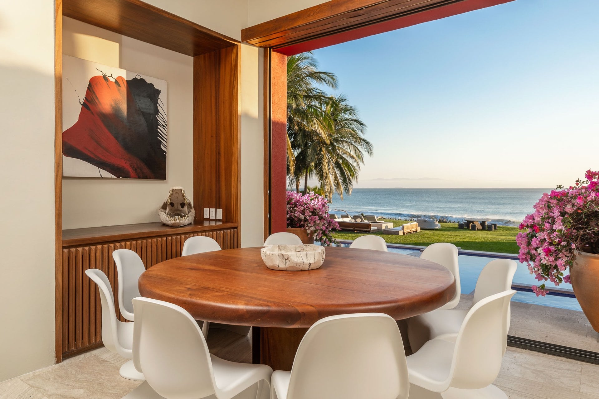 A round walnut-slab dining table seats eight on white tulip chairs; an abstract painting hangs on the wood-paneled wall while bougainvillea and the Pacific fill the open frame behind, Villa Pacífica, Ranchos Estates, Punta Mita.