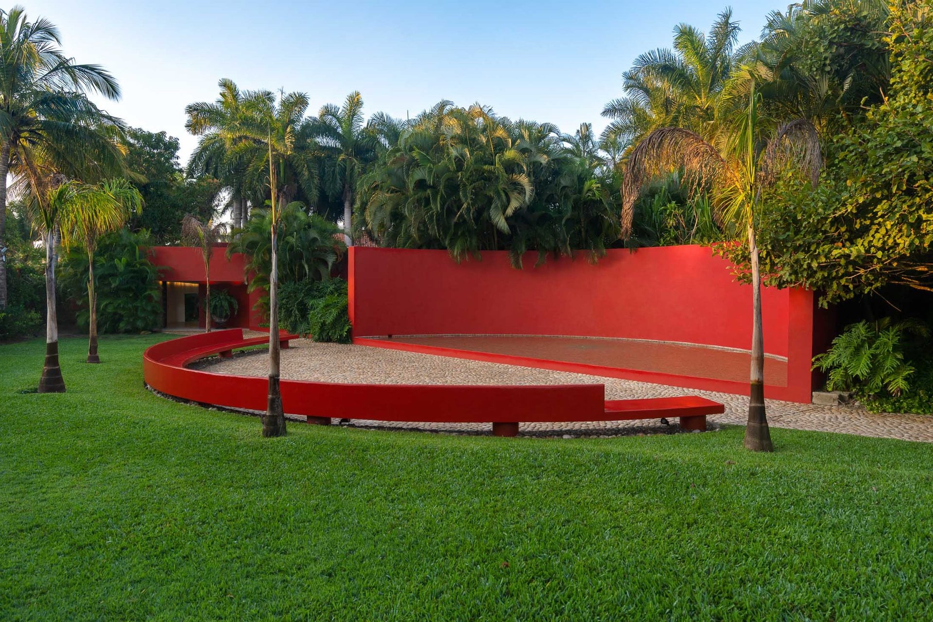 A curved low red-painted wall spirals across the lawn forming an amphitheater seating area, royal palms rising behind it against a blue-hour sky, Villa Pacífica, Ranchos Estates, Punta Mita.