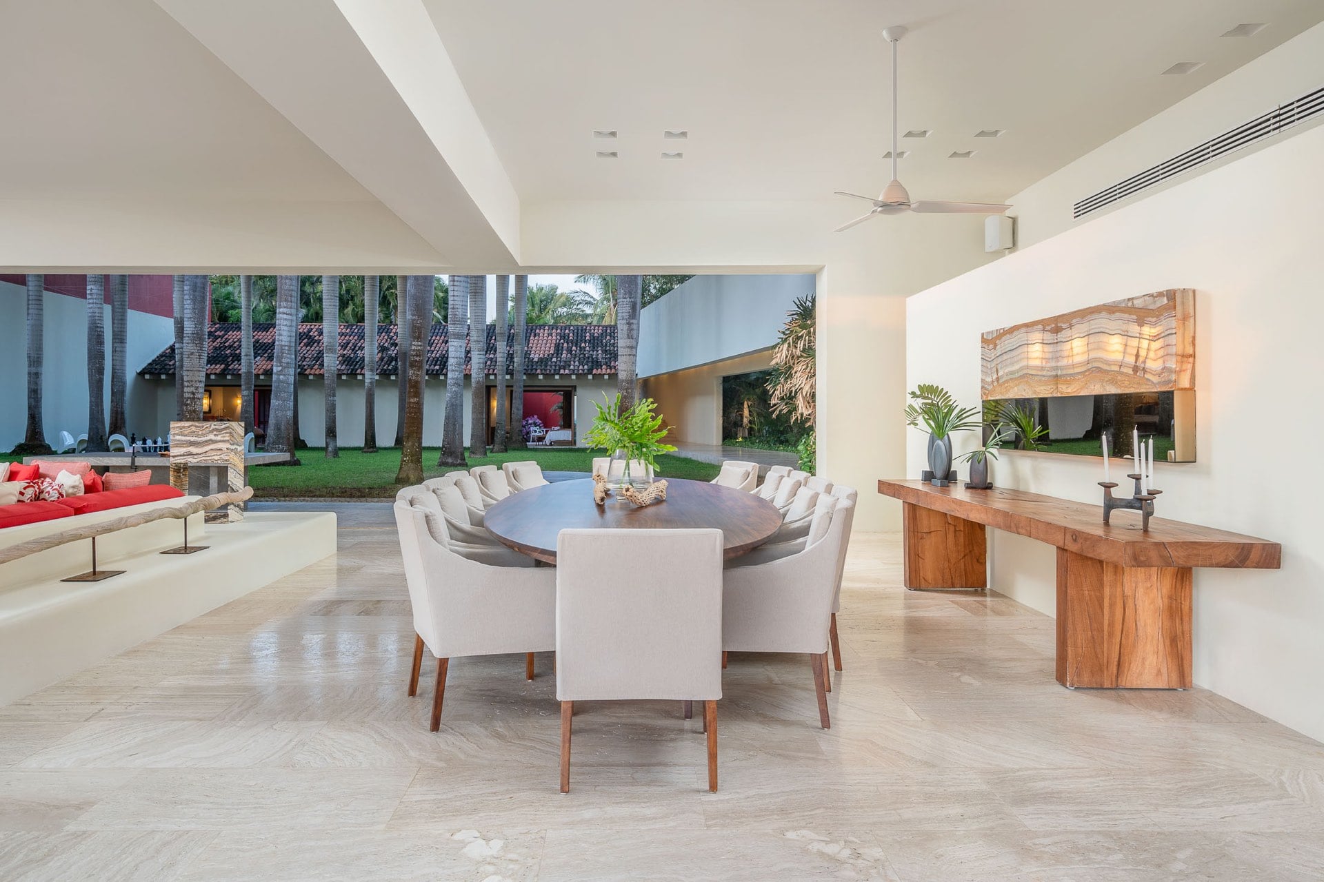 The covered dining pavilion seats twelve around a large round table; a backlit onyx slab panel hangs over the wooden console while royal palms and the tiled roof of the estate fill the open garden wall, Villa Pacífica, Ranchos Estates, Punta Mita.