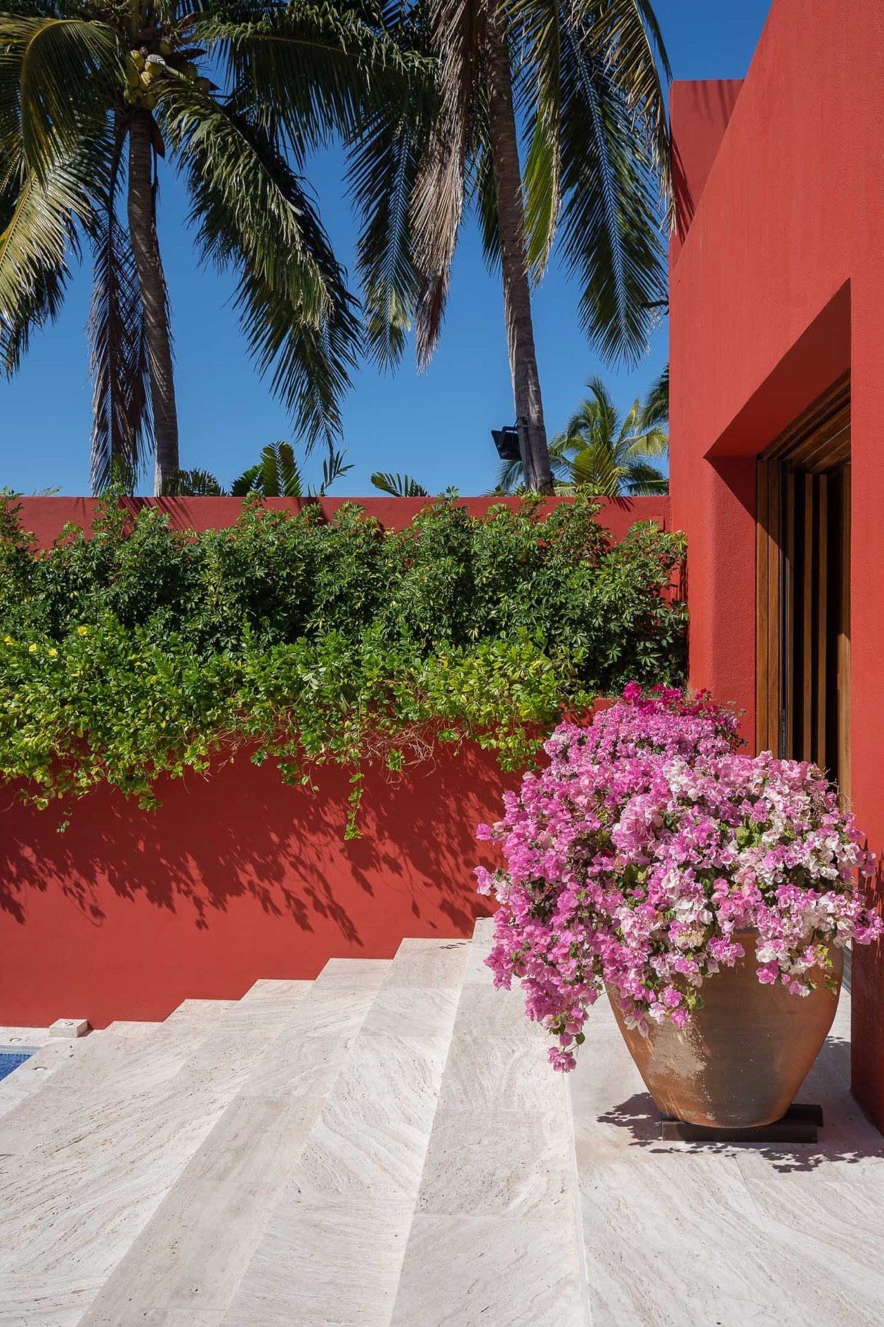 A large terracotta pot overflowing with pink bougainvillea sits on pale travertine steps at the villa entrance, the red perimeter wall draped with trailing greenery against a clear blue sky, Villa Pacífica, Ranchos Estates, Punta Mita.