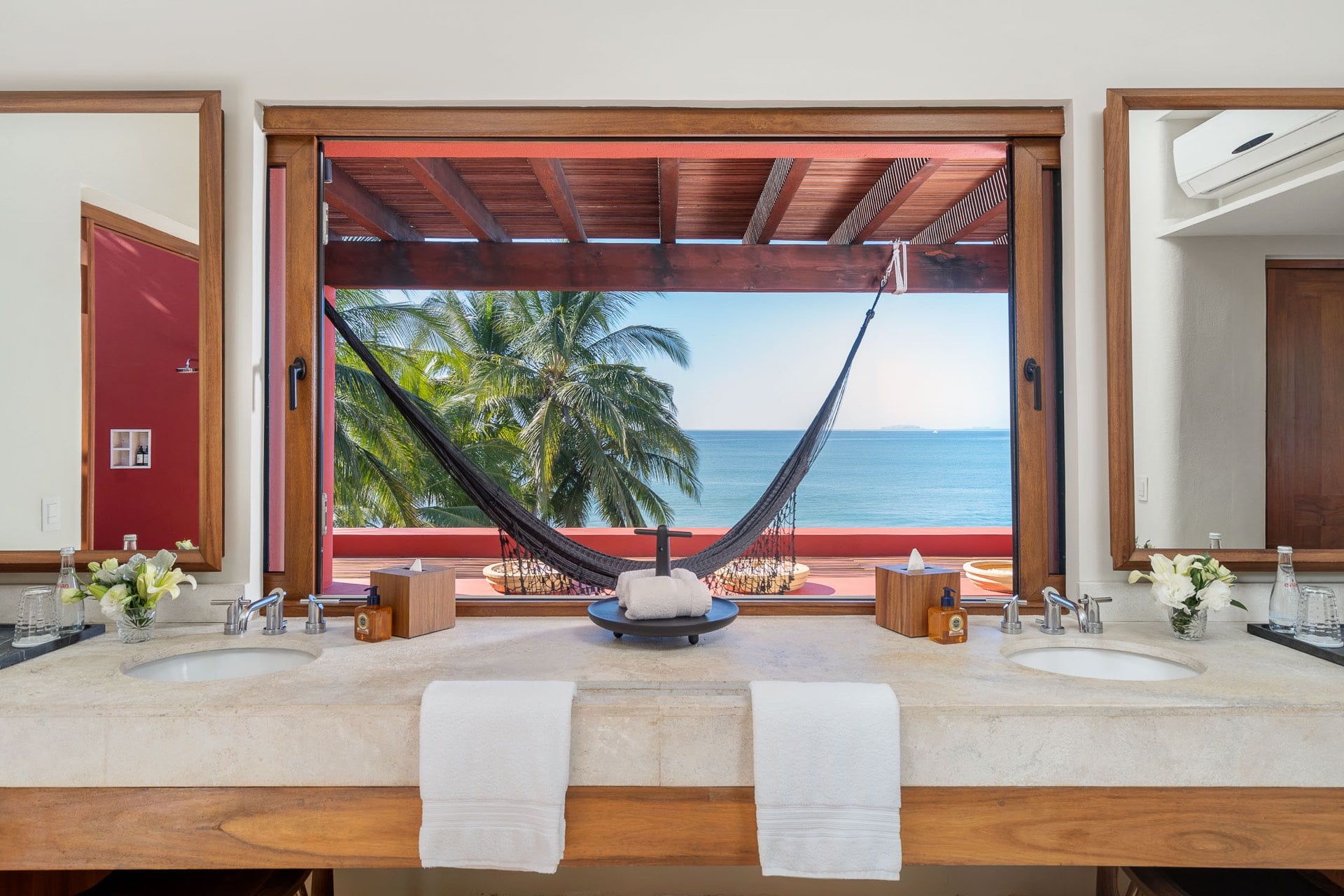 A dual-sink marble vanity faces a wide picture window that frames a hammock on the terrace pergola with the Pacific and palms behind it, Villa Pacífica, Ranchos Estates, Punta Mita.