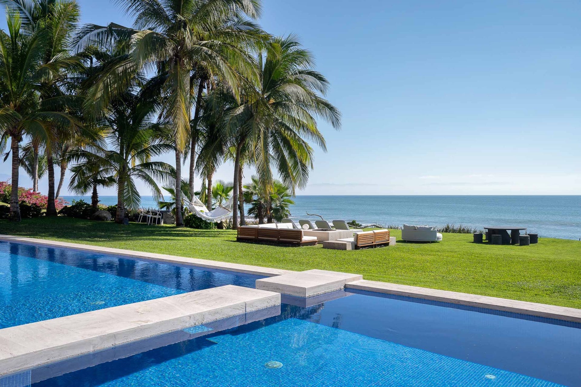 Two pool levels step down through a broad lawn toward the beach, loungers and a daybed sofa arranged on the grass while palms frame the Pacific under a clear midday sky, Villa Pacífica, Ranchos Estates, Punta Mita.