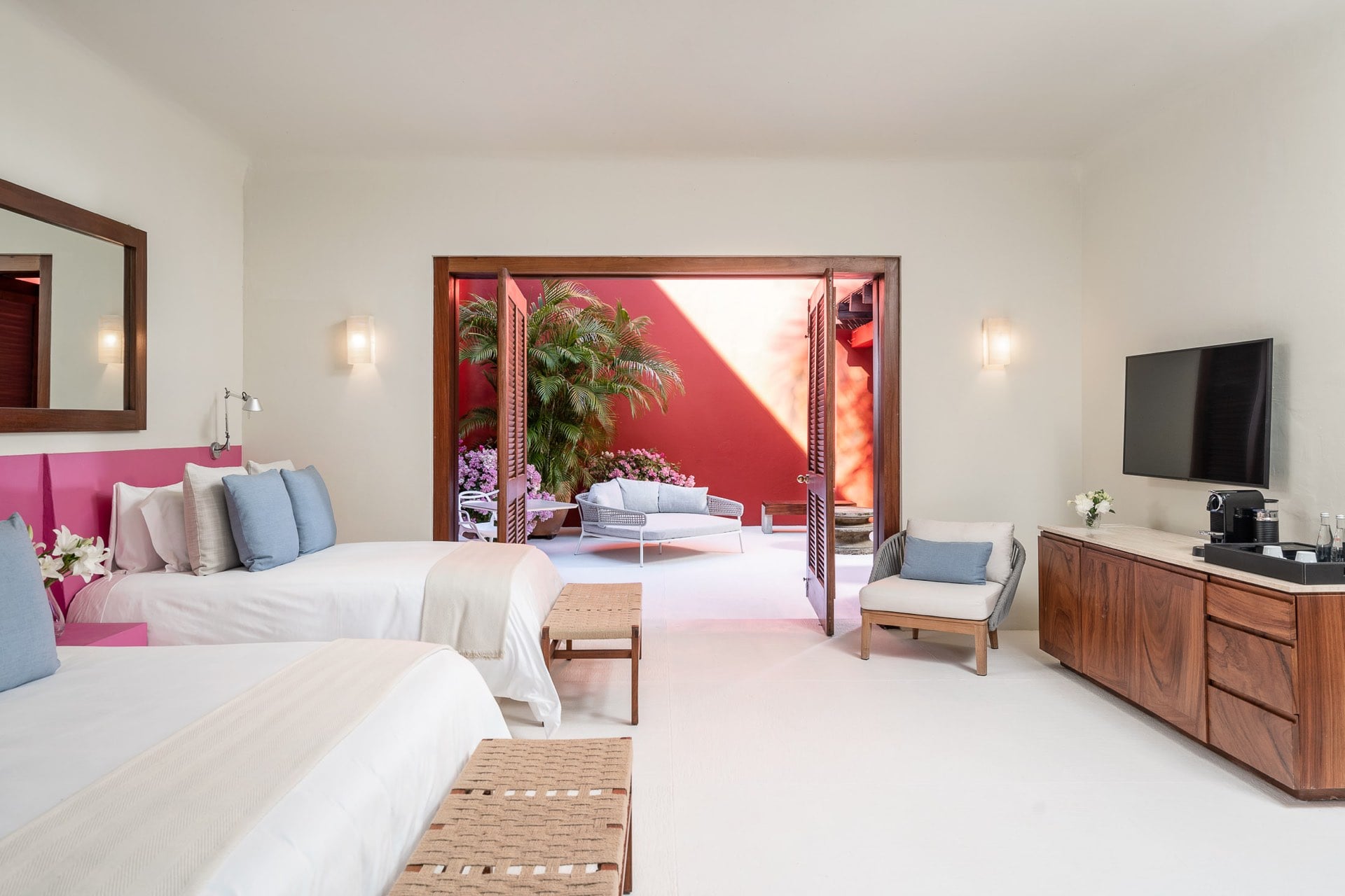 Two twin beds with pink upholstered headboards face French doors that open onto a private courtyard filled with bougainvillea and palms lit by afternoon sun, Villa Pacífica, Ranchos Estates, Punta Mita.