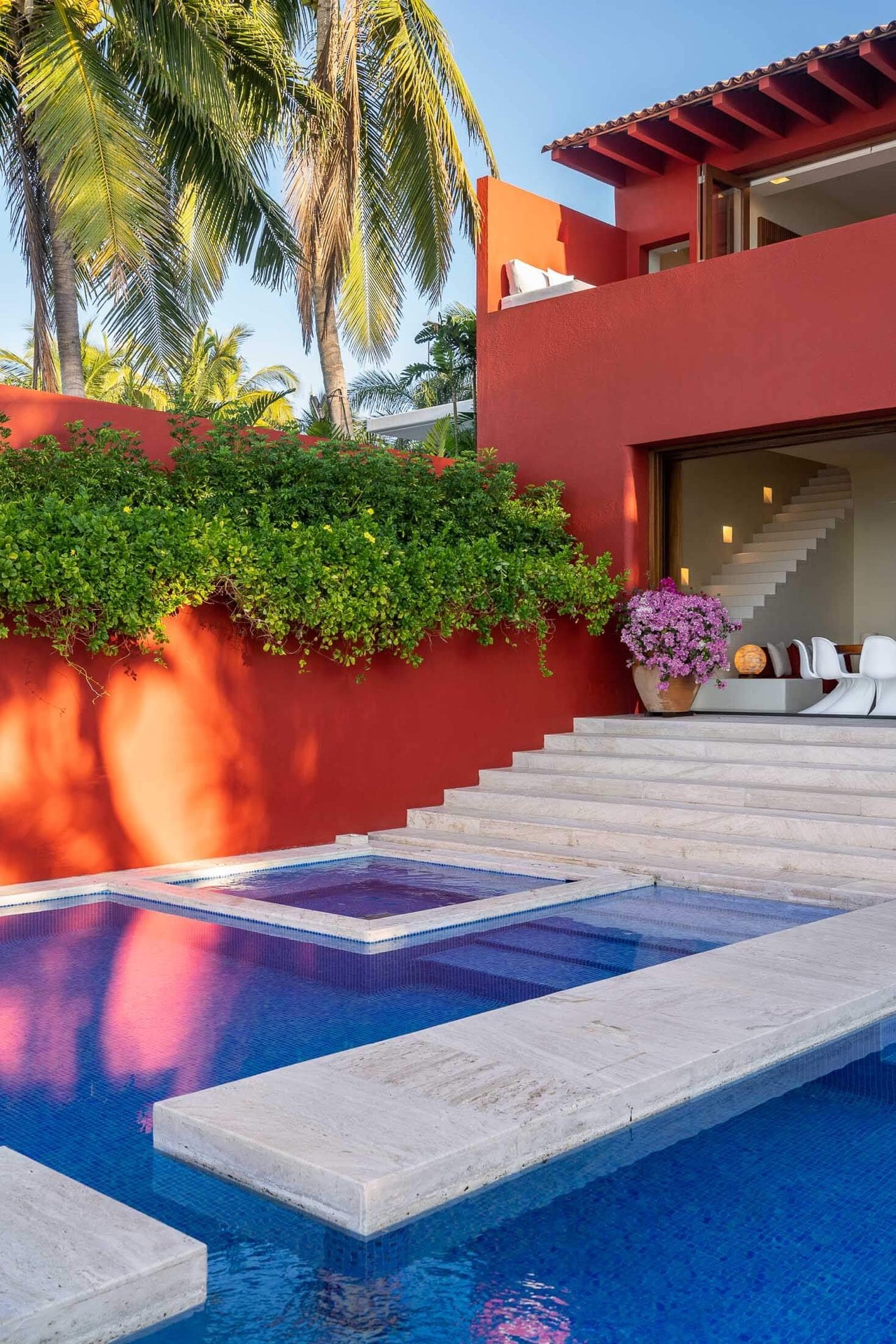 The pool and recessed hot tub sit at the base of broad travertine stairs, the red villa wall rising behind them while palms and bougainvillea soften the afternoon light, Villa Pacífica, Ranchos Estates, Punta Mita.