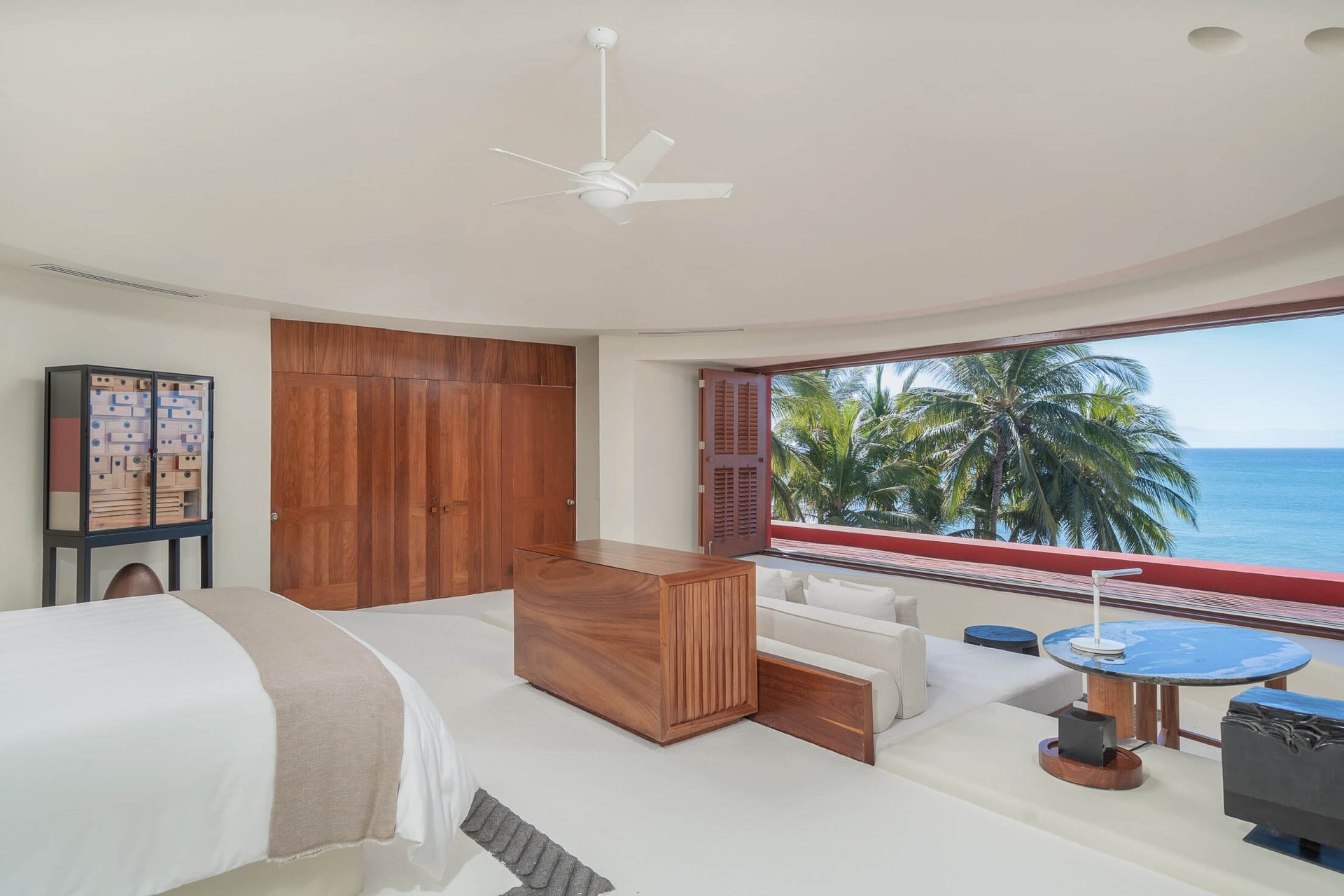 The primary suite’s panoramic shuttered window opens wide to palms and open ocean, a sitting area with a blue table and walnut wardrobe wall occupying the foreground, Villa Pacífica, Ranchos Estates, Punta Mita.