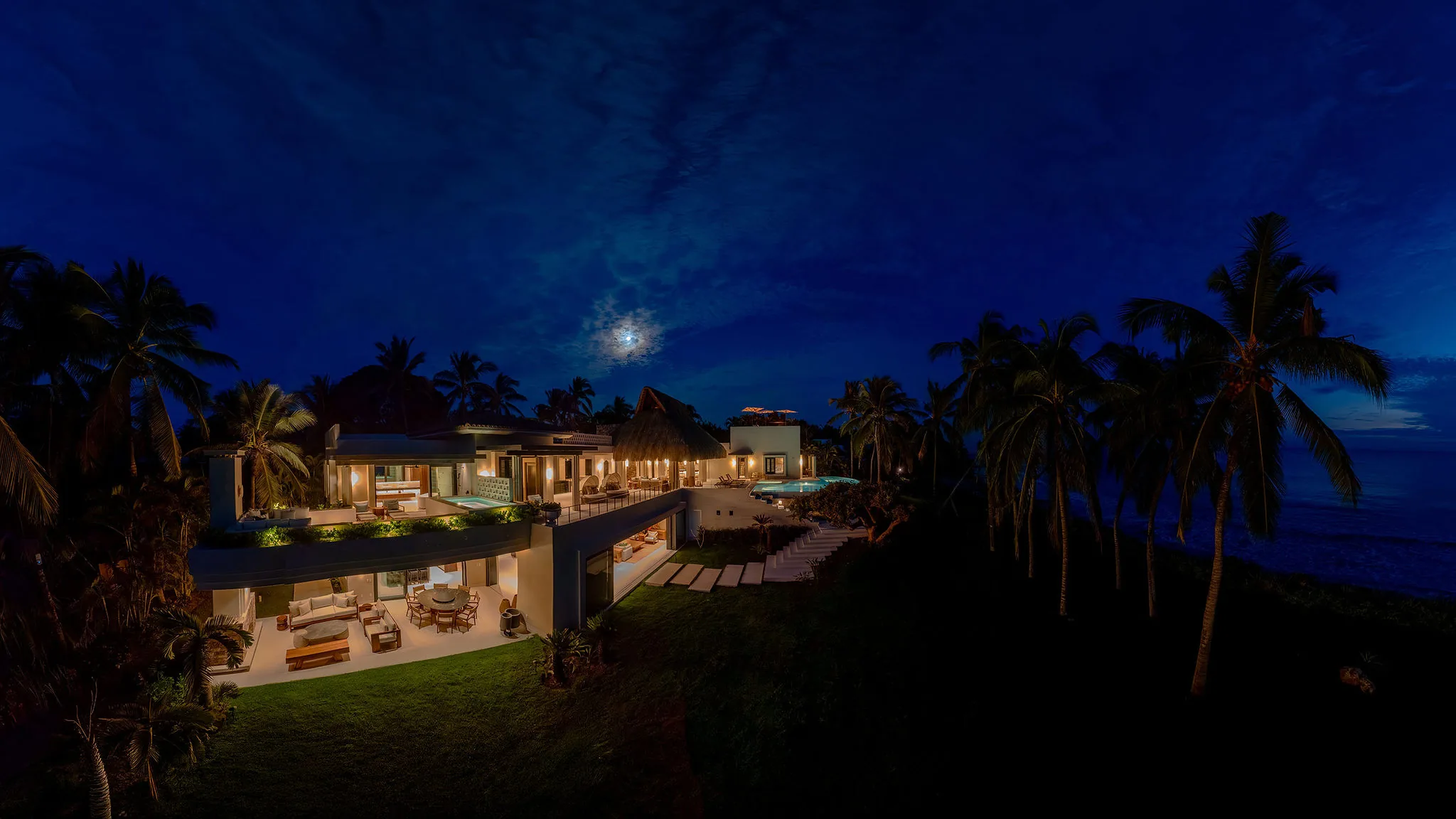 Casa Happy Paws, a luxurious modern villa in Ranchos Estates, Punta Mita, shines warmly at night by the beach and palm trees.