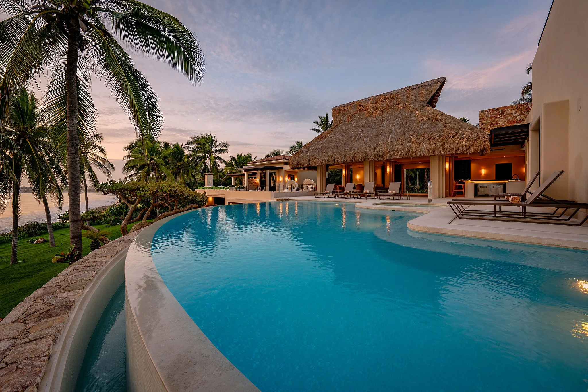 Casa Happy Paws in Punta Mita’s Ranchos Estates: luxury villa, thatched roof, infinity pool, ocean views, lush gardens at sunset.