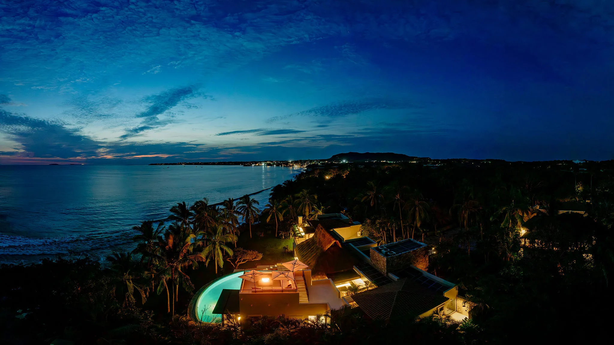 Ranchos Estates Punta Mita: Palm-lined resort and pool lit at dusk, ocean views near Casa Happy Paws, glowing shoreline lights.