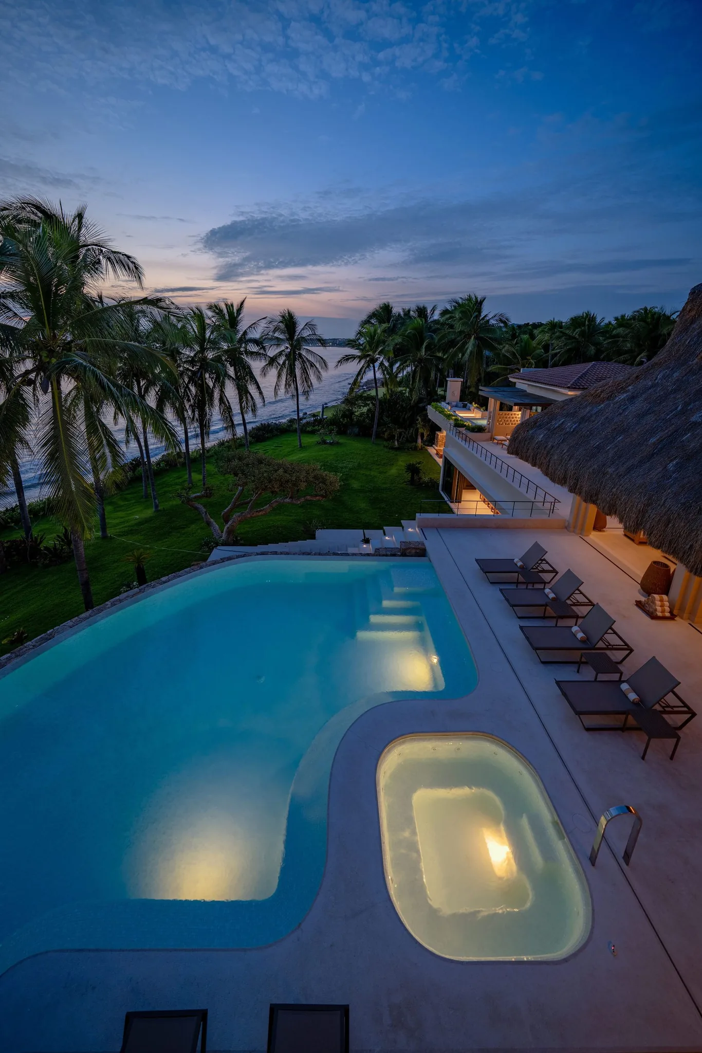 Sunset view of lit pool and hot tub at Casa Happy Paws, Punta Mita, Ranchos Estates; palm trees, thatched-roof, beach.