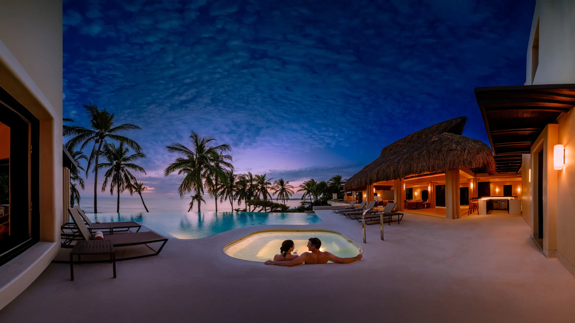 Couple unwinds in hot tub at Casa Happy Paws, Ranchos Estates, Punta Mita, with palm trees and infinity pool at sunset.